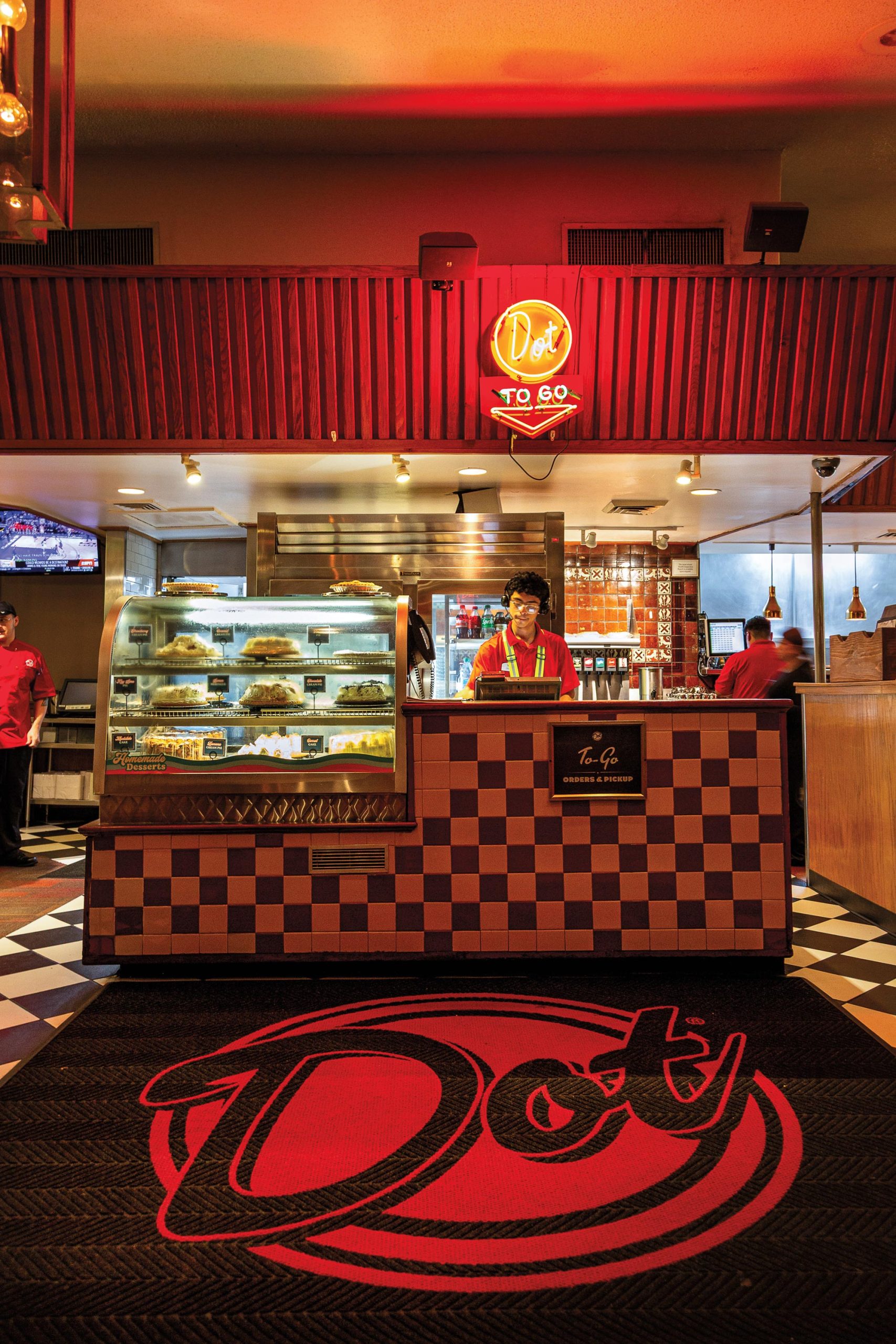 A restaurant worker in a red shirt and green suspenders behind a checkerboard-tiled counter under a neon sign that reads Dot To-Go