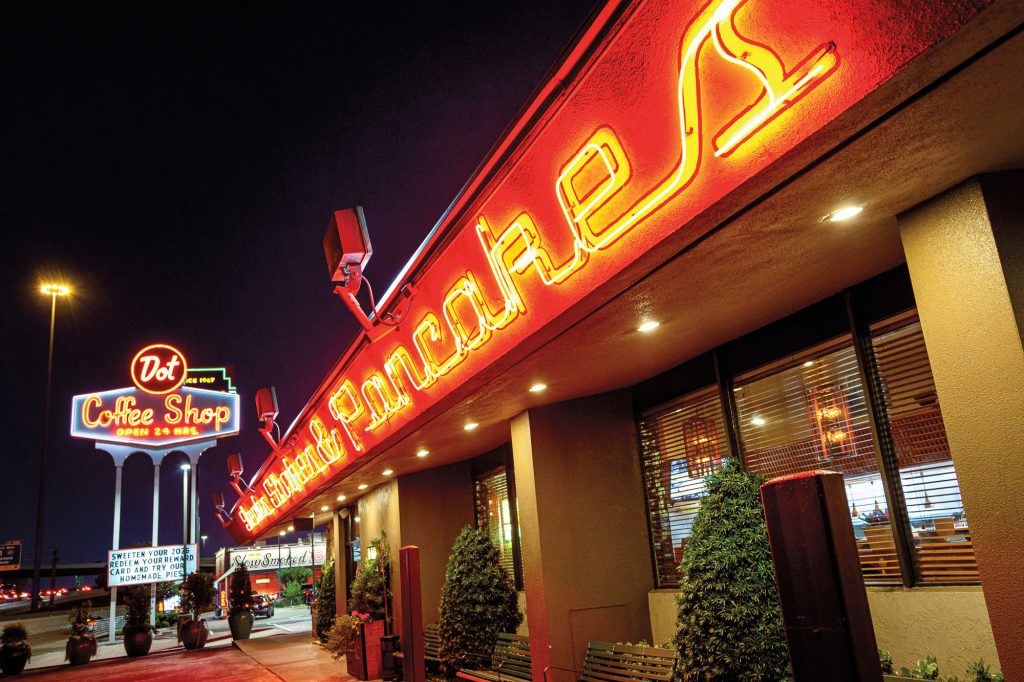 A neon sign at night that reads Dot Coffee Shop Open 24 Hrs. next to an awning with neon lettering that reads Steaks, Shakes & Pancakes