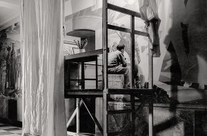 An artist painting while sitting on a wooden platform