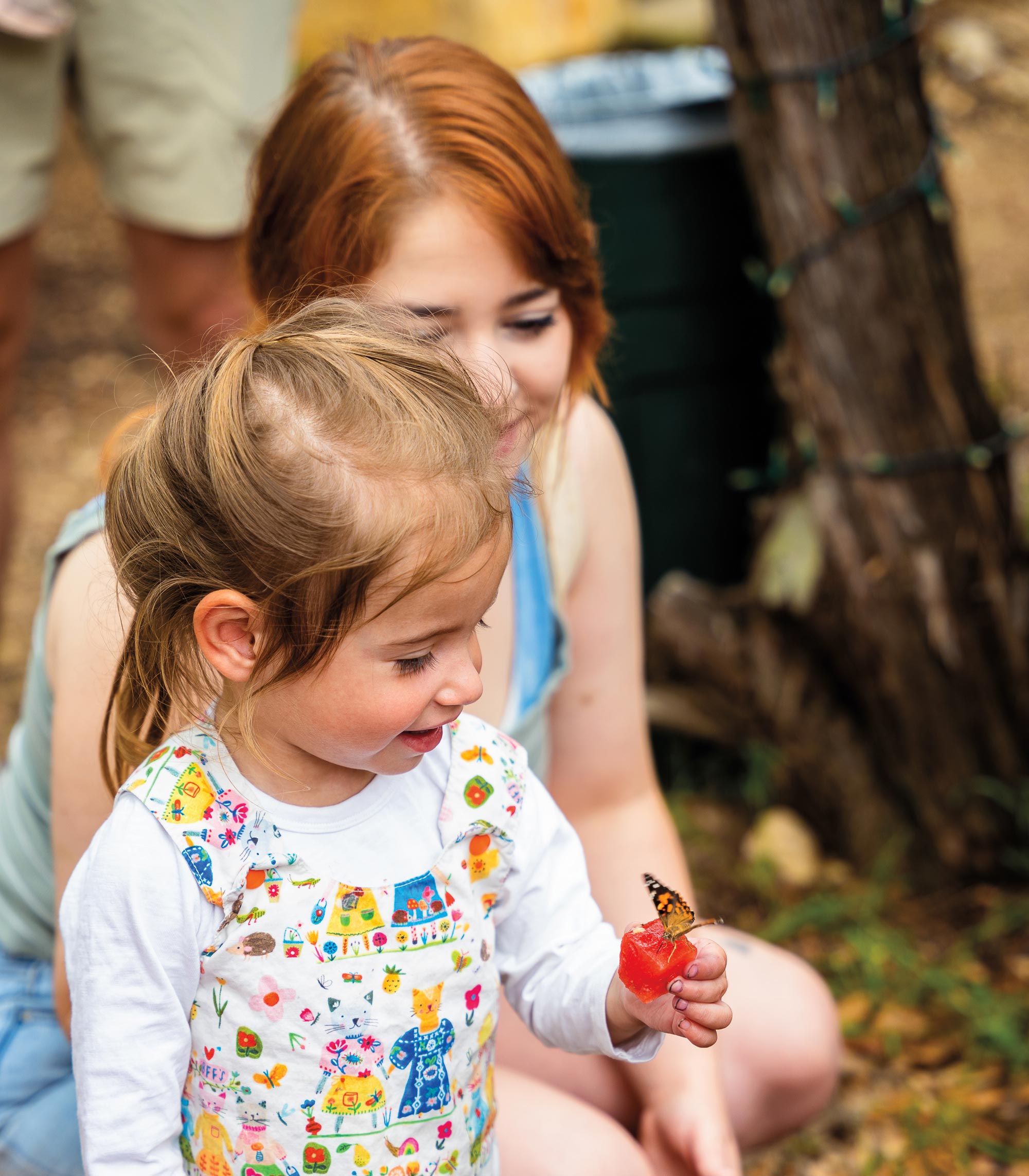 A woman crouching behind a small girl who is holding a piece of red fruit with a butterfly on top of it