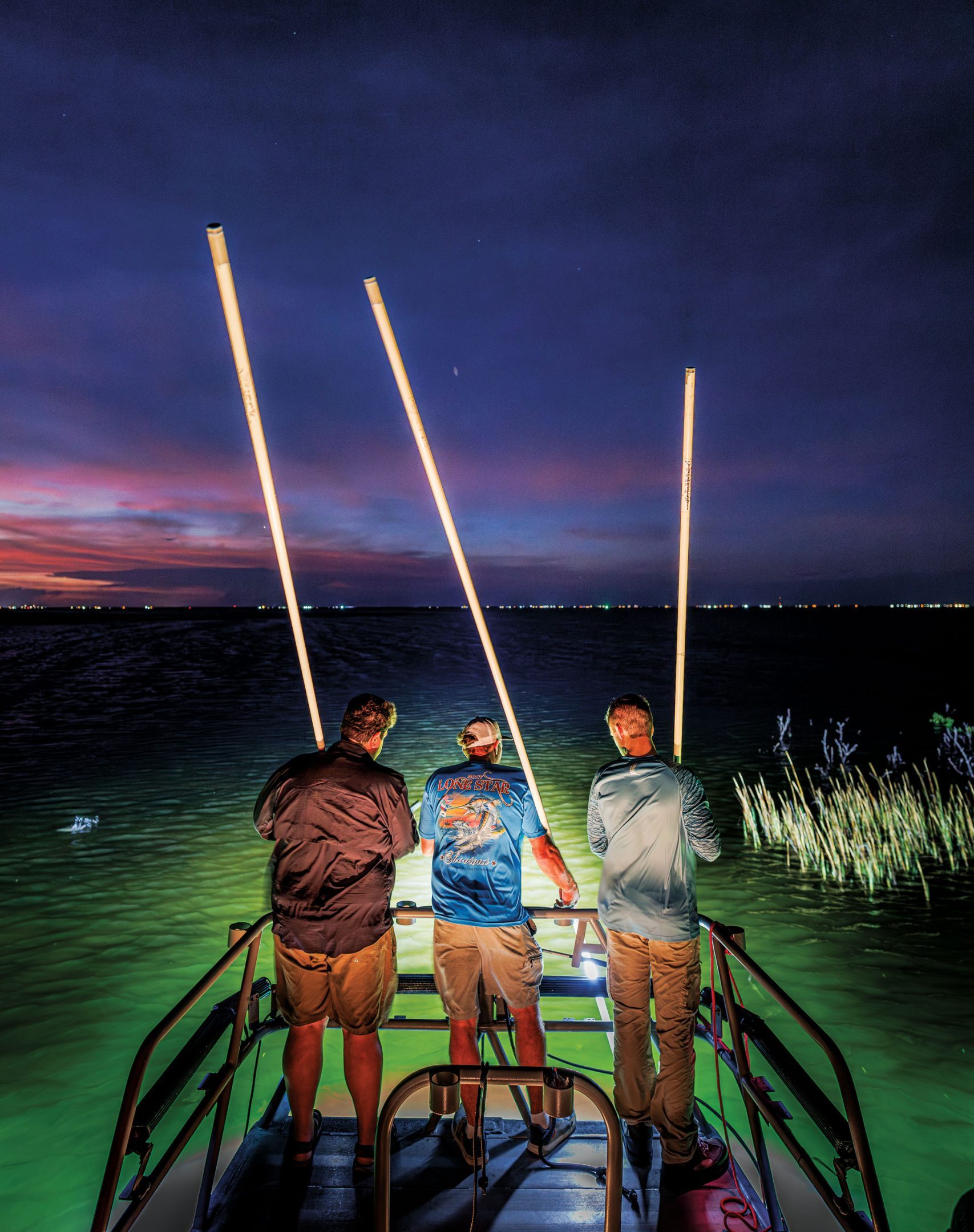 Three men stand on the edge of a boat with rods in their hands ready to spear something in illuminated waters at night