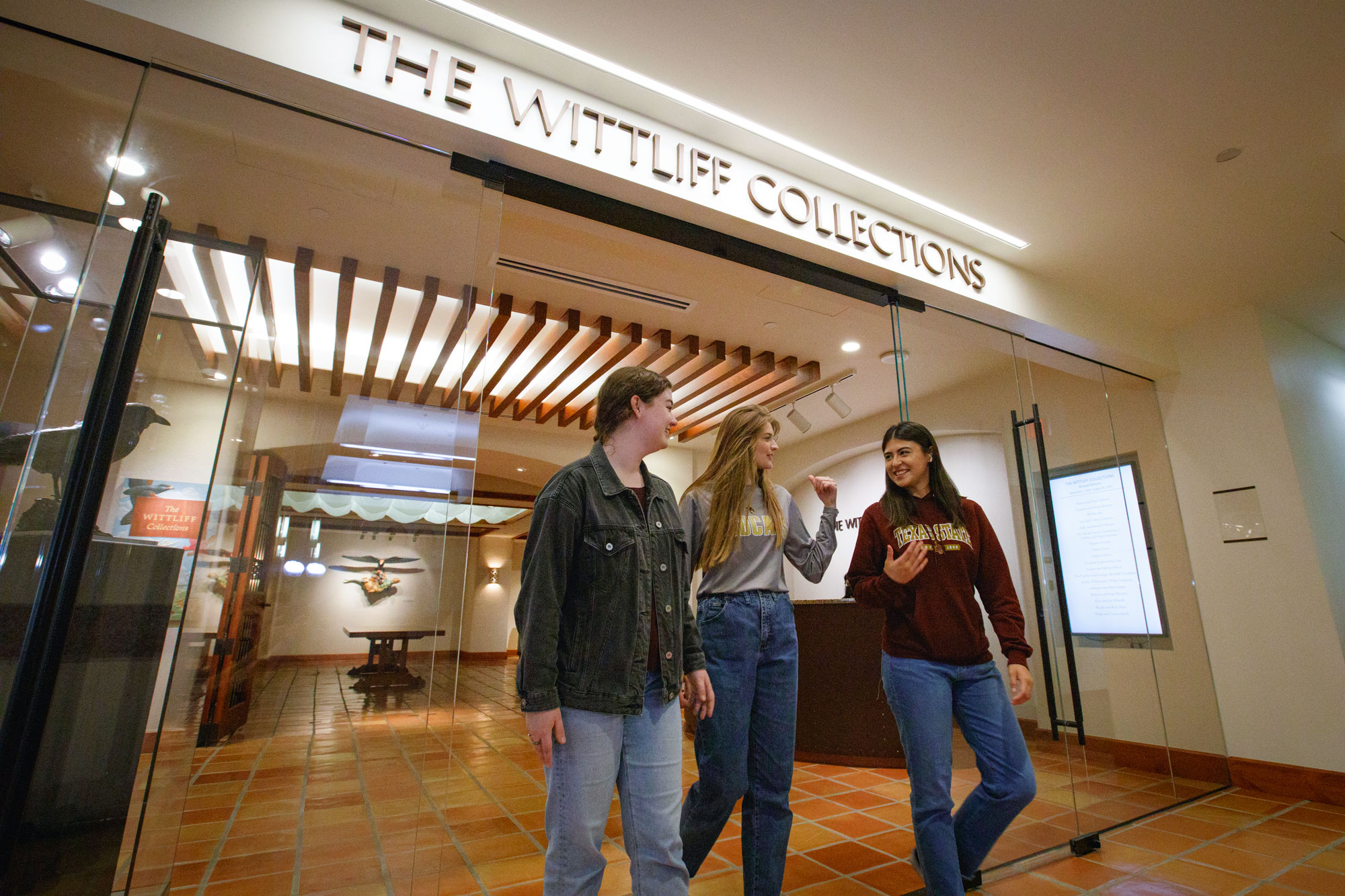 Three young women exit a museum entrance with a sign that reads "The Wittliff Collections"