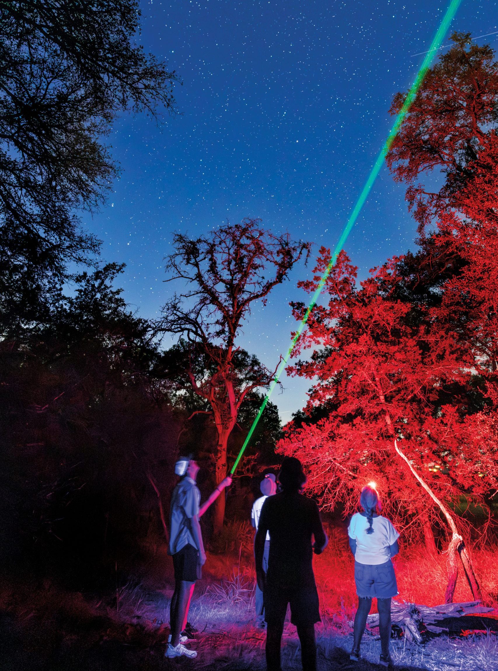 People in a forested park at night hold green lasers up to the sky