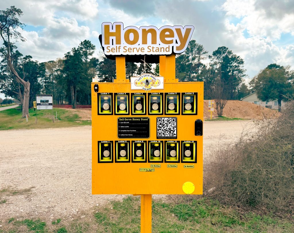 A yellow honey vending machine outdoors that reads Honey Self Serve Stand with bottles of honey inside and a QR code for payment