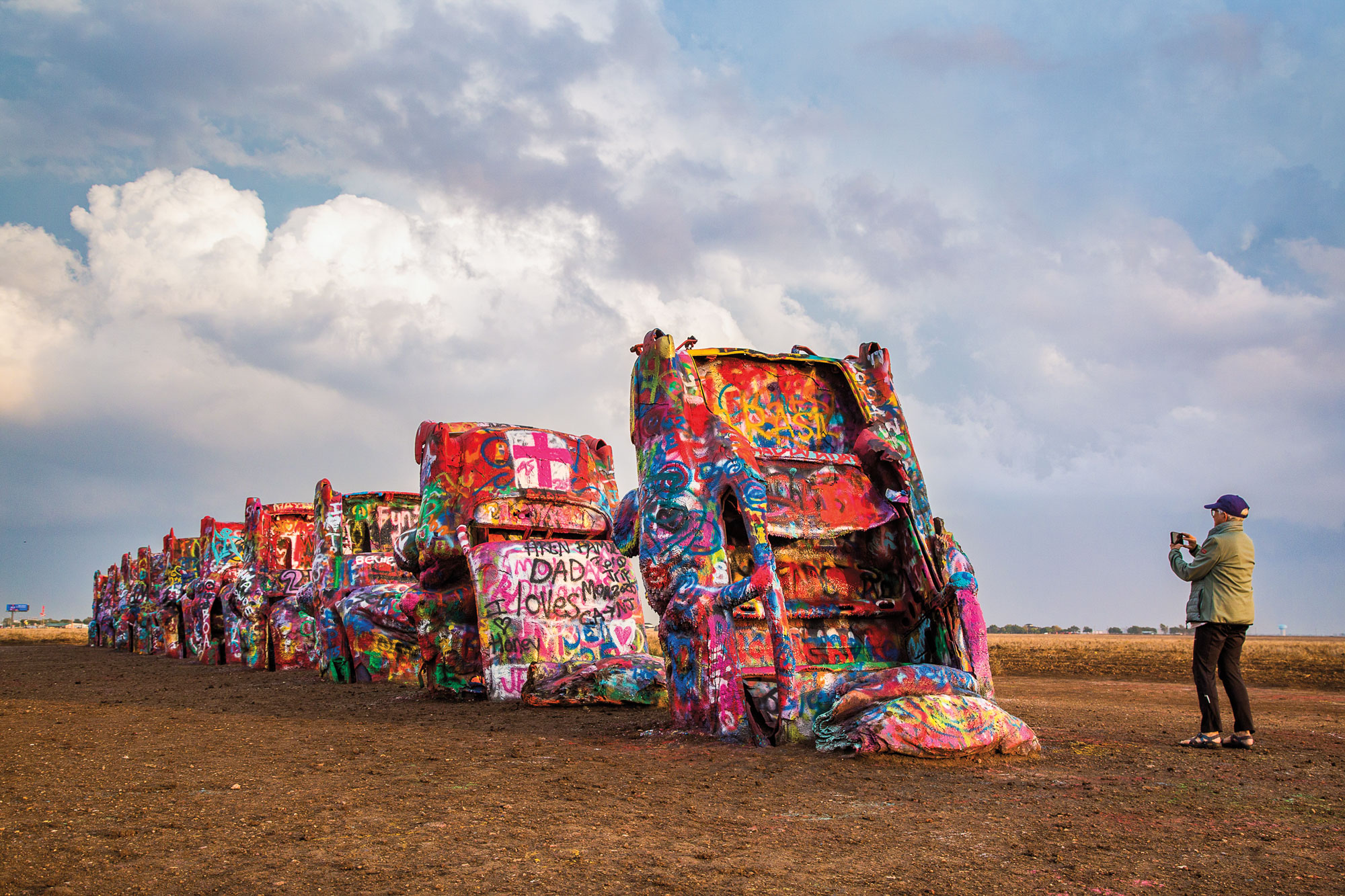 Several old Cadillacs covered in colorful graffiti sit in the dirt