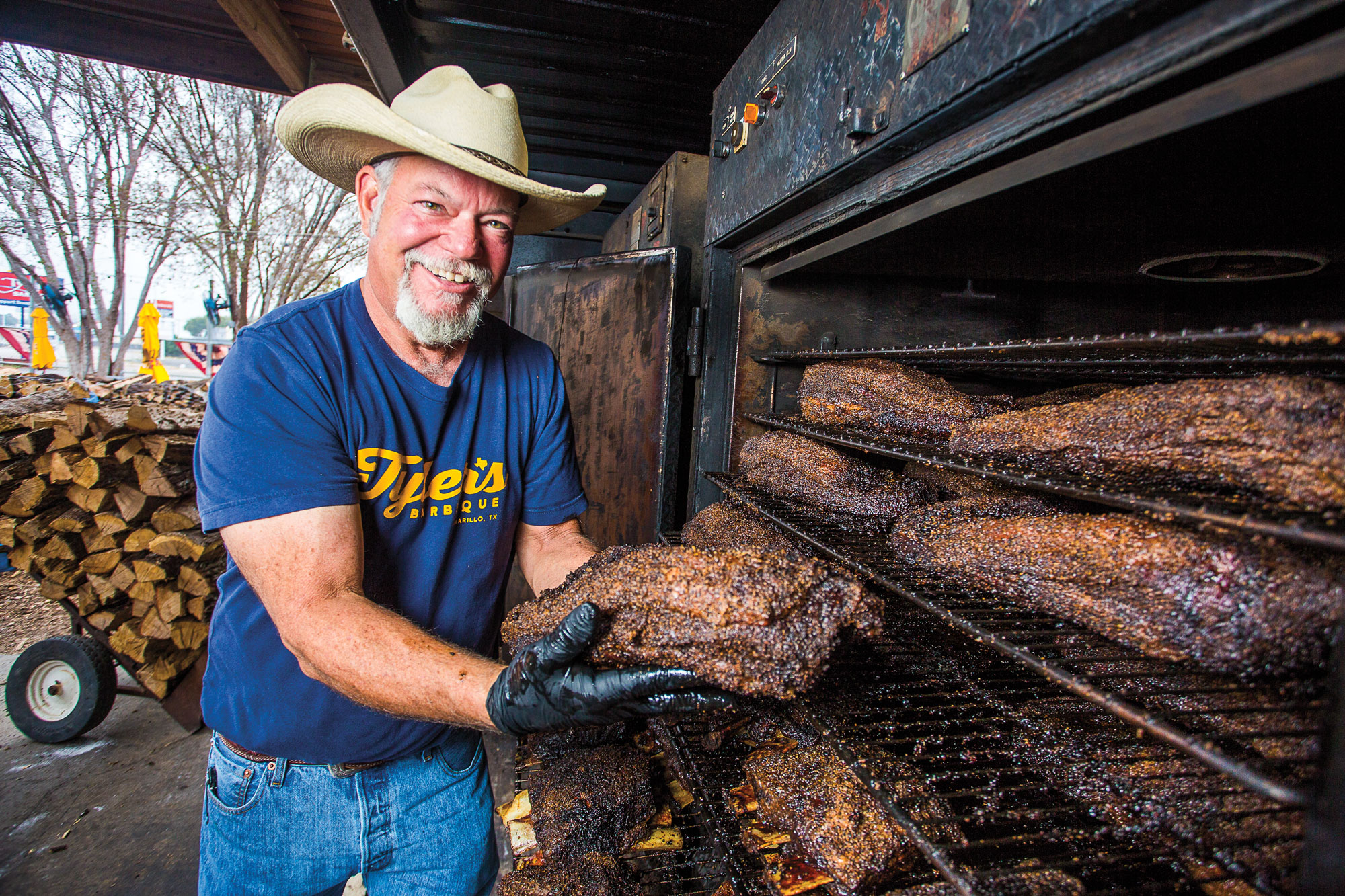 An older white man with a white beard in a cowboy hat stands in front of a large smoker while smiling and holding a brisket
