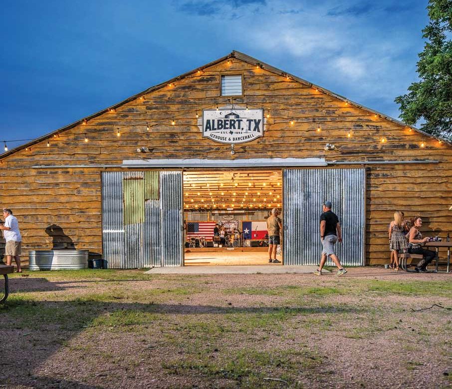 A barnhouse style dance hall illuminated at dusk with its doors open and people comingin and out. There's a sign on the front that reads "Albert, TX"