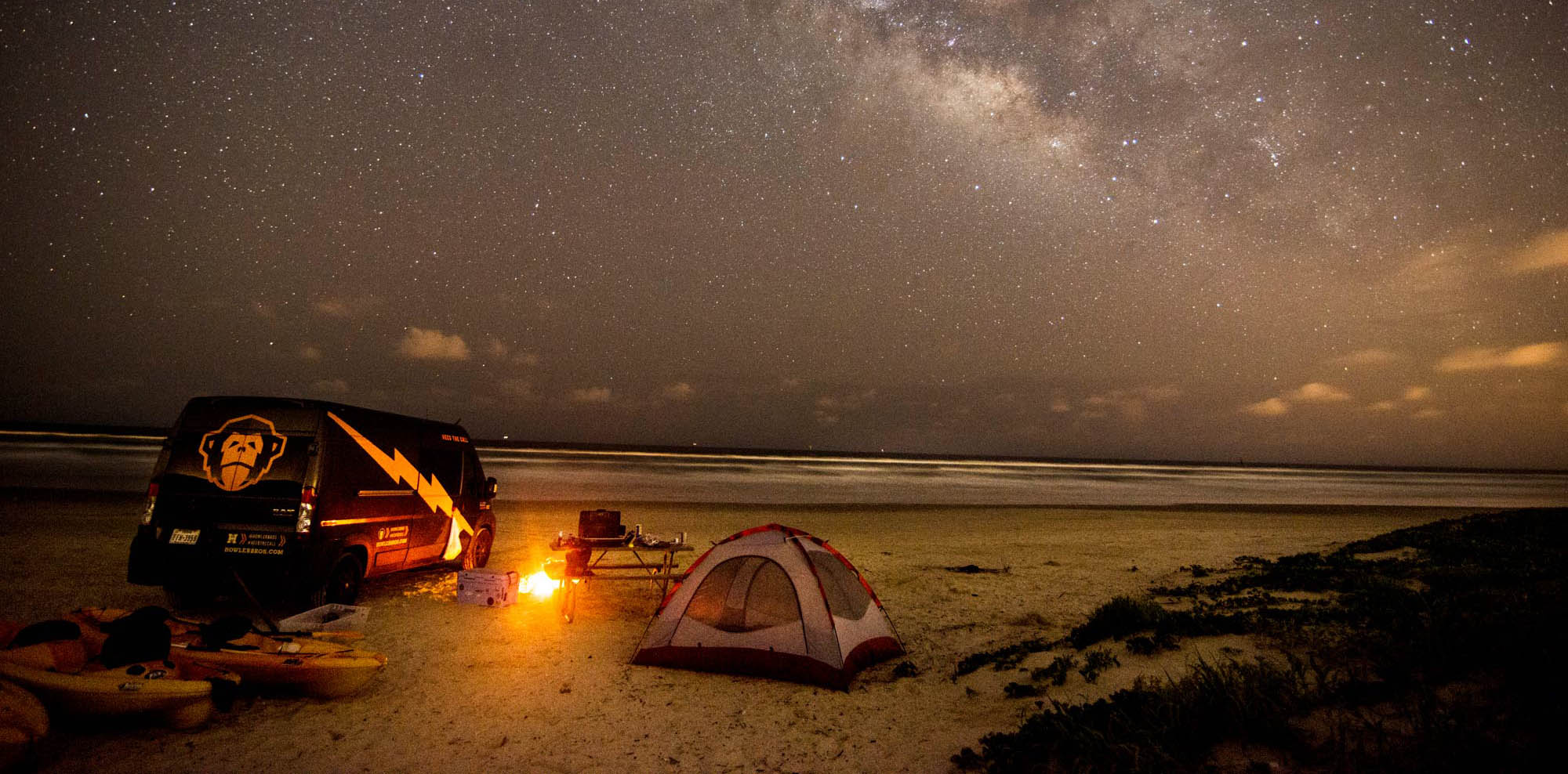 A black van with a yellow illustration of a monkey's head and a lightning bolt next to a campfire, cooler, table, and tent under the stars on the beach