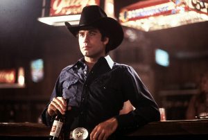 A man in a Western shirt tucked into pants with a big belt buckle holding a bottle of beer and wearing a black cowboy hat
