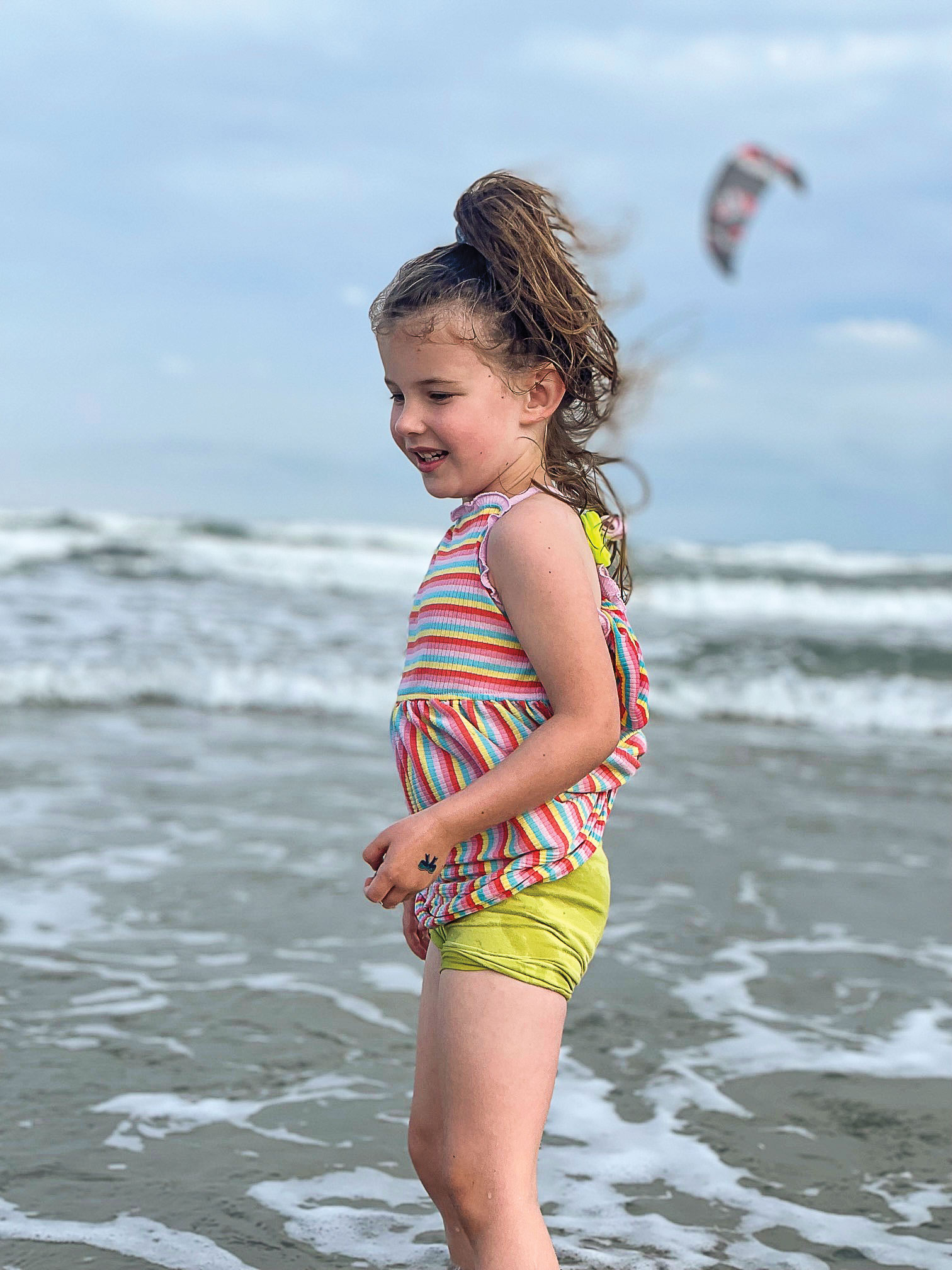 A young little girl with a ponytail in a swimsuit stands in the ocean.