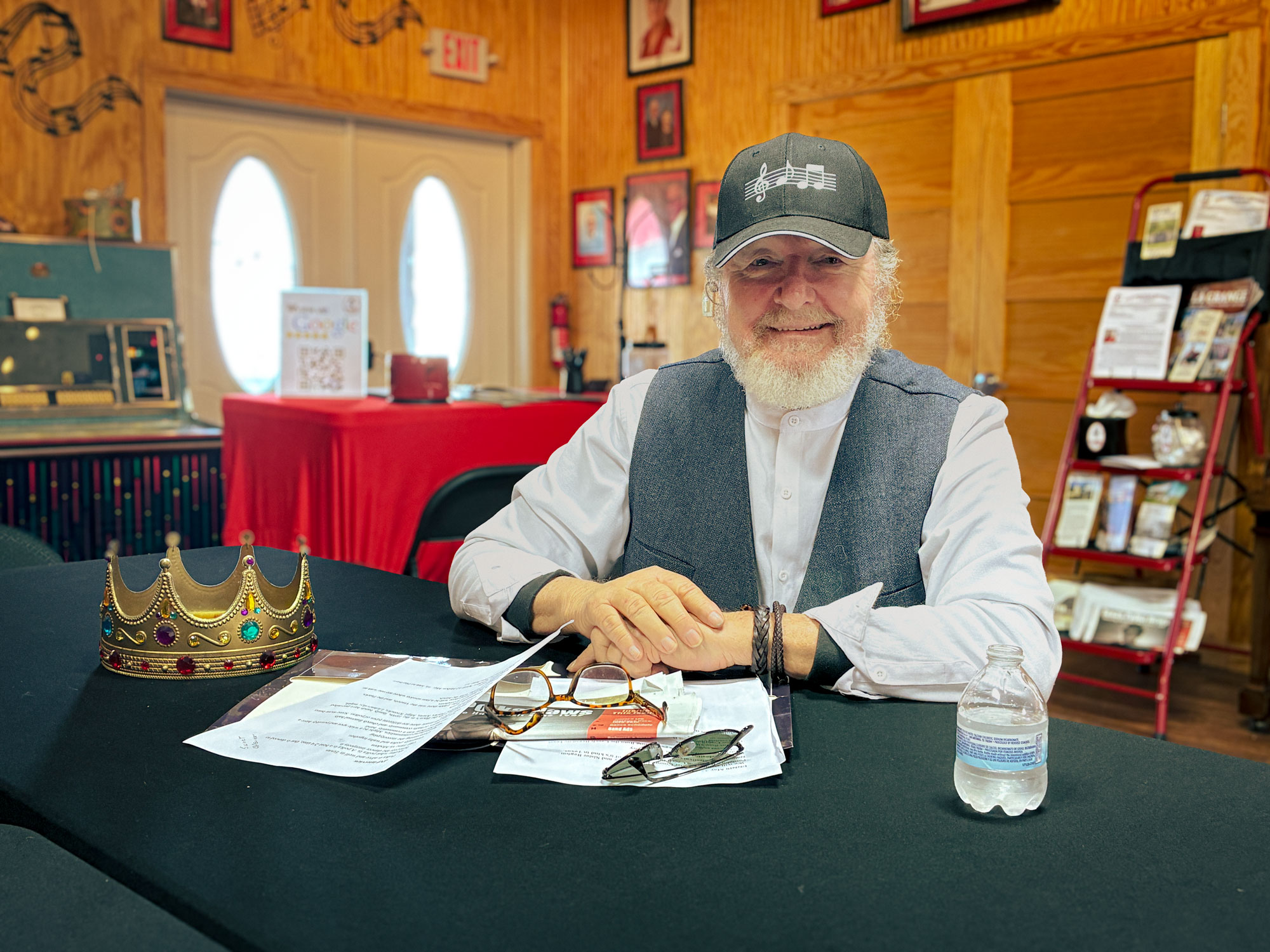 An older man in a cap sits a table inside a wooden building.
