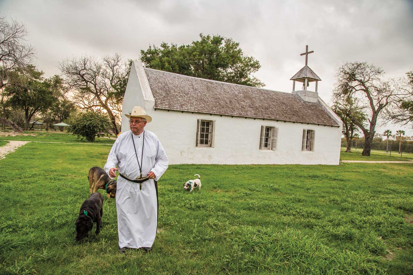 A man in a white robe and cowboy hat stands with two dogs in front of a small white chapel