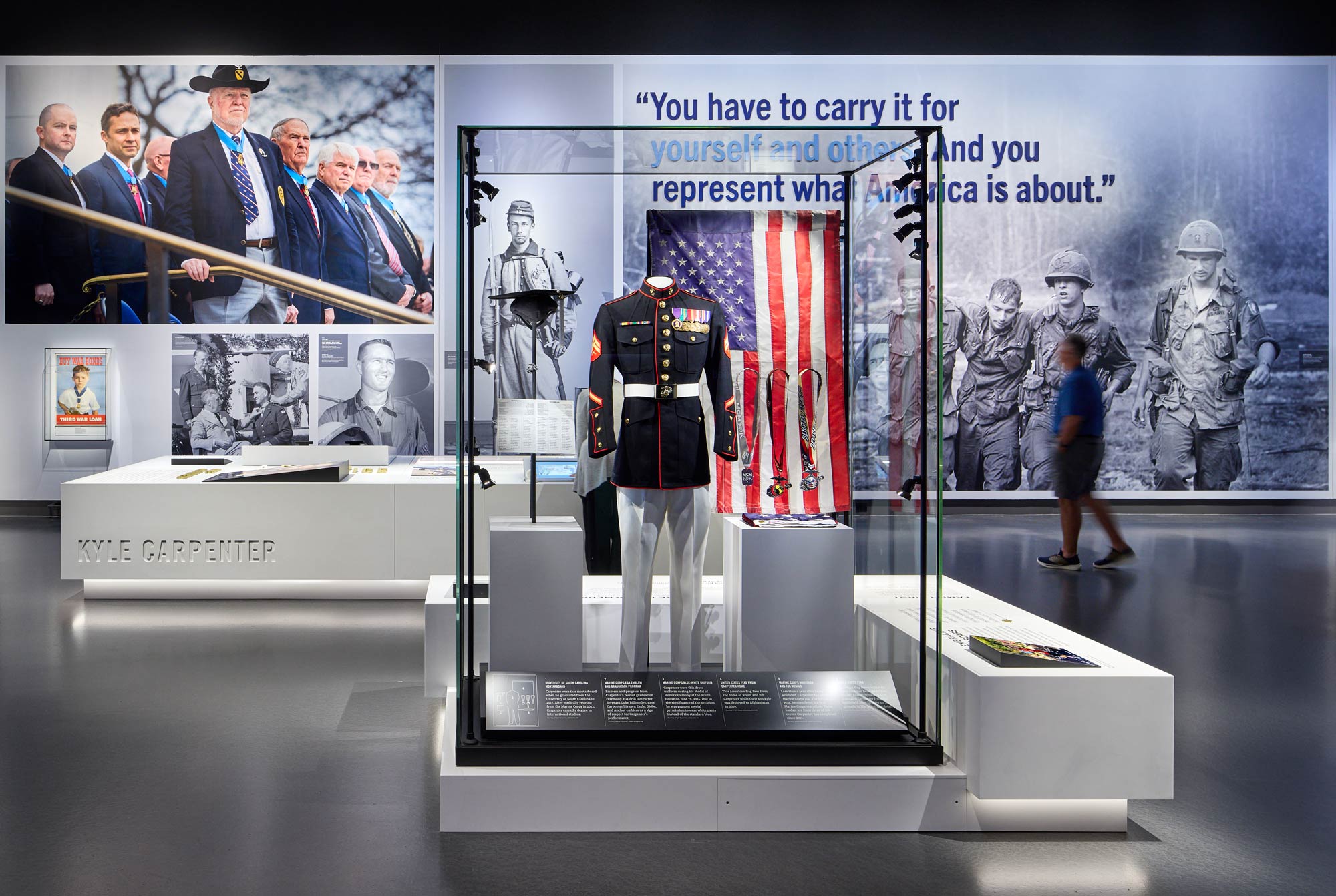 A glass case containing a solider's uniform on a mannequin next to several medals with a man walking behind and to the right of it