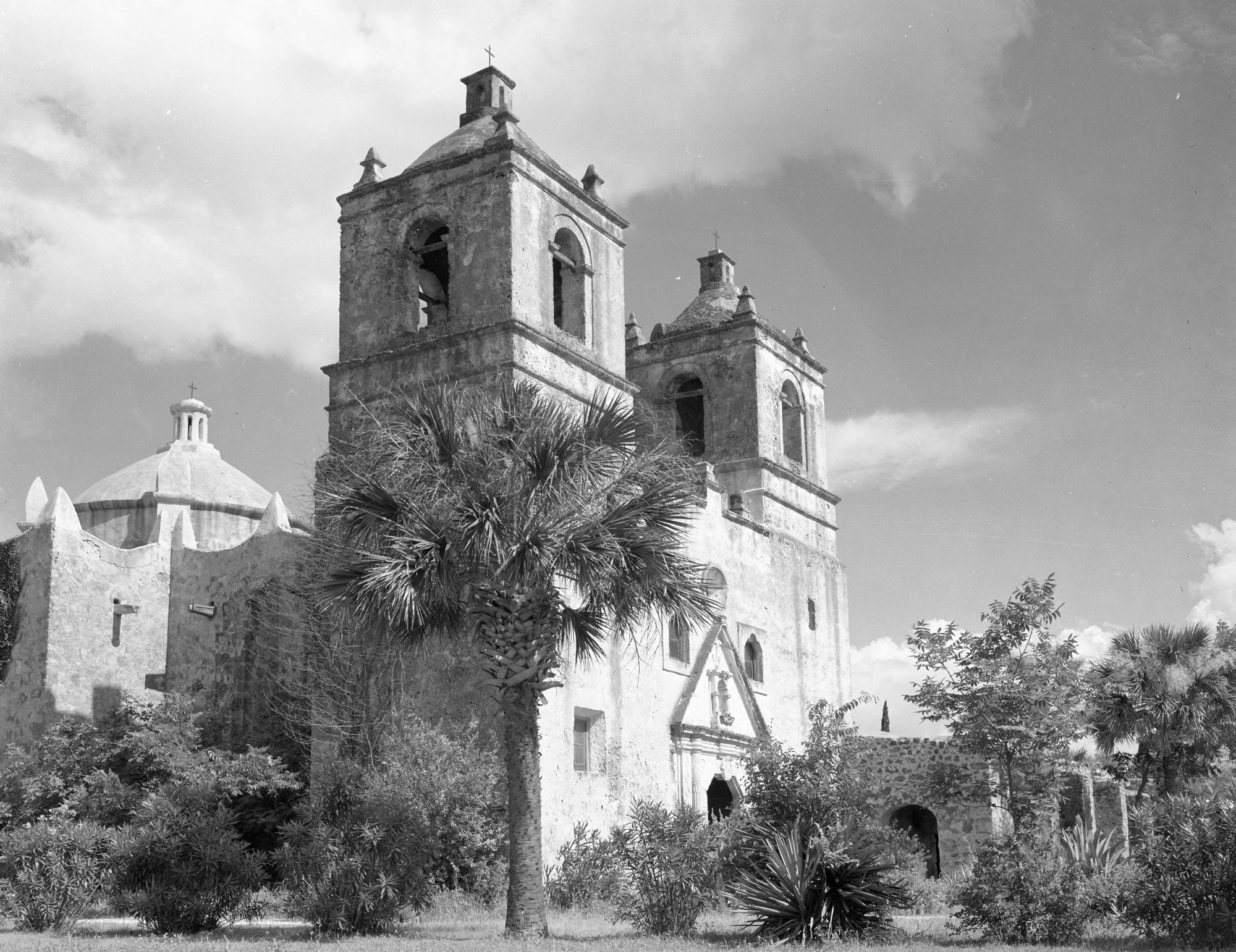 A large stone structure with multiple towers and a dome behind palm trees and bushes