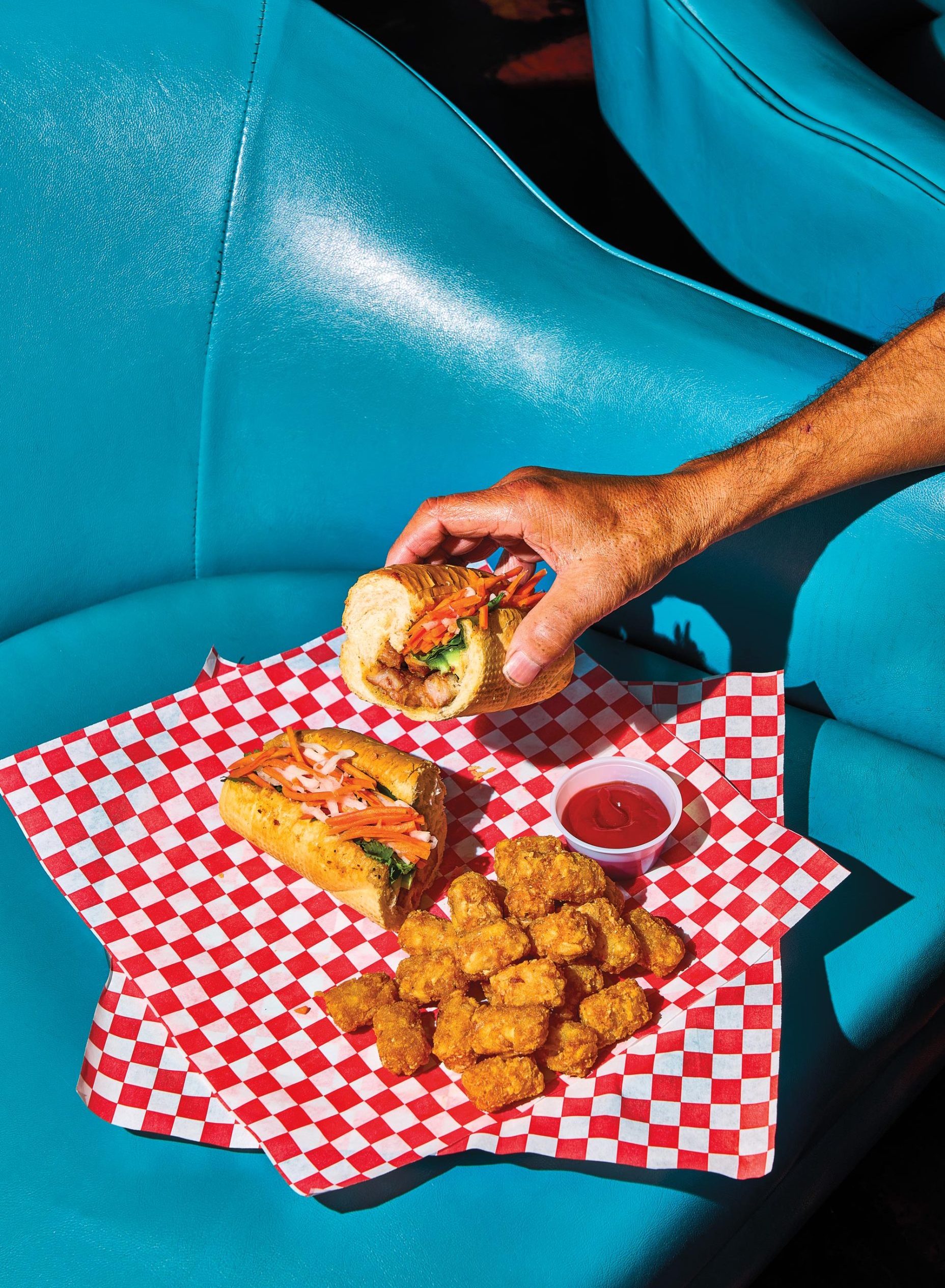 An outstretched arm holding half of a bahn mi sandwich over two checkered napkins containing the other half of the sandwich, tater tots, and a plastic ramekin of ketchup