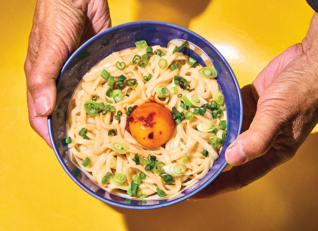 Two hands holding a blue bowl filled with noodles and scallions topped by an egg yolk