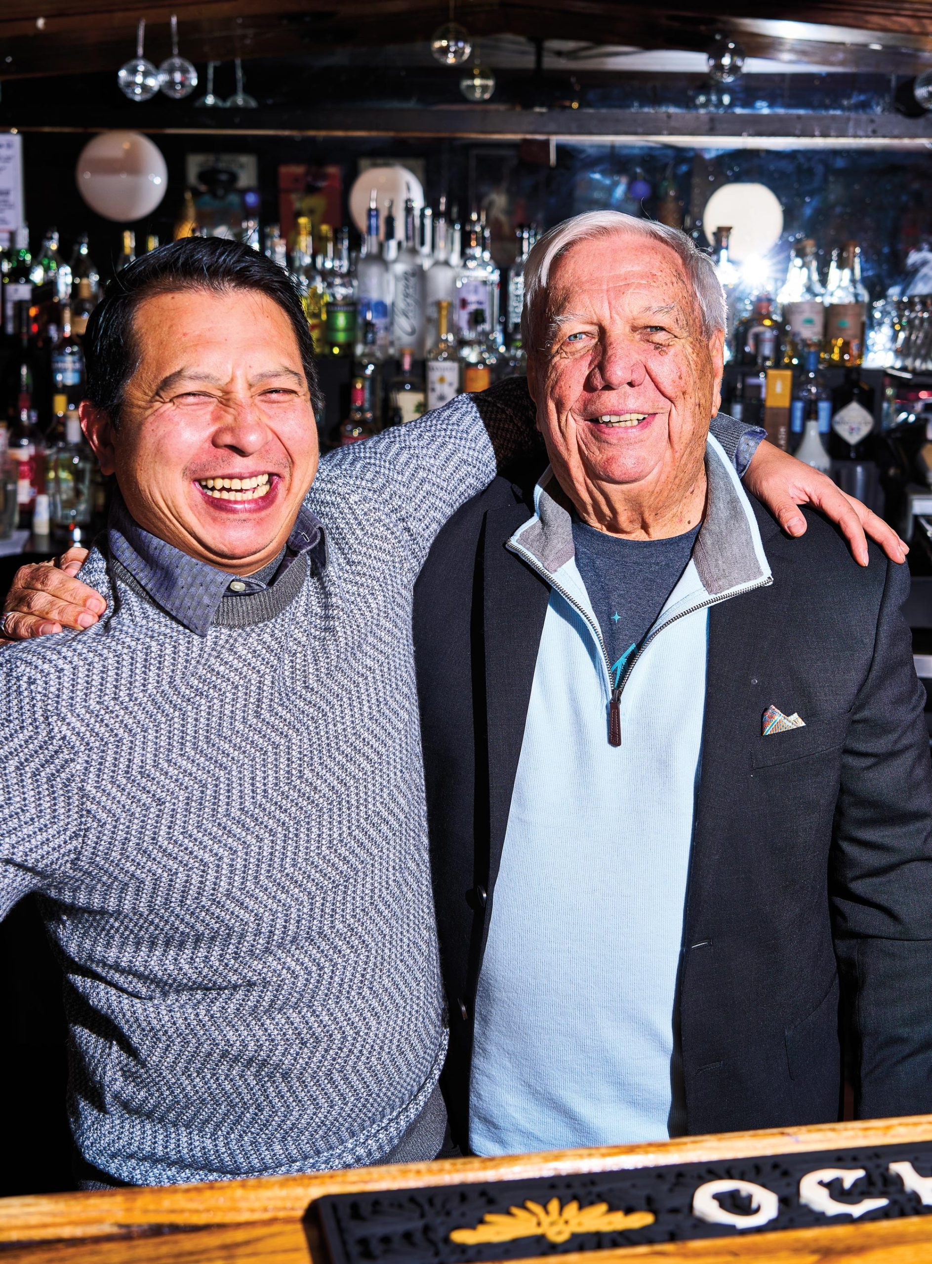 Two men, the younger of whom is on the left of the image, with their arms around each other and smiling behind a bar