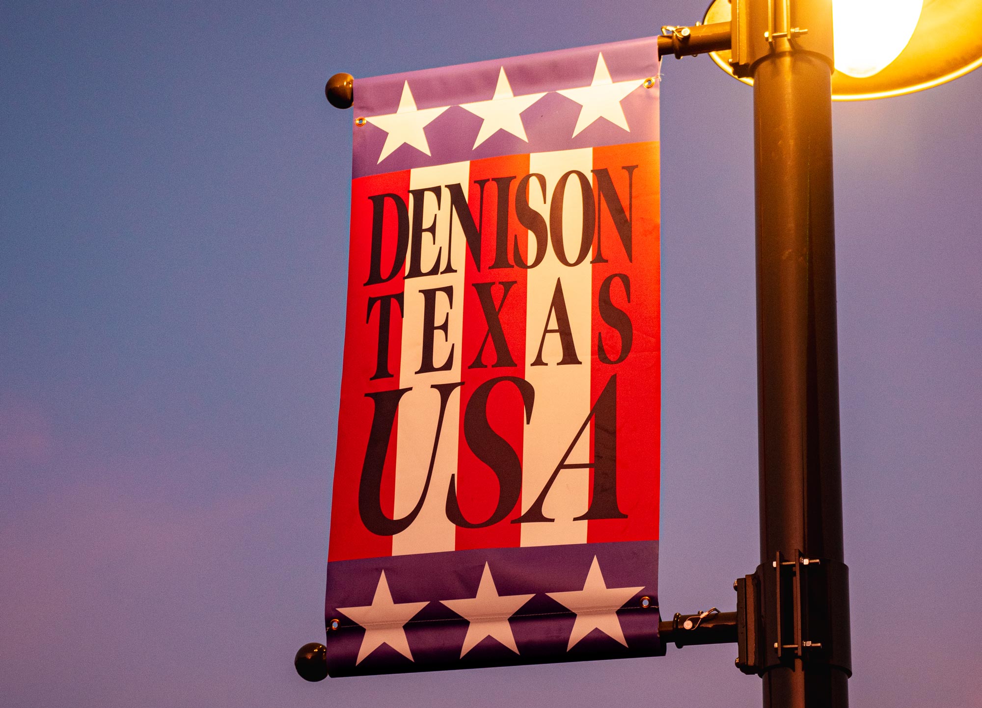 A red, white, and blue flag with six stars and the words DENISON TEXAS USA printed on it hanging from a lit lightpole