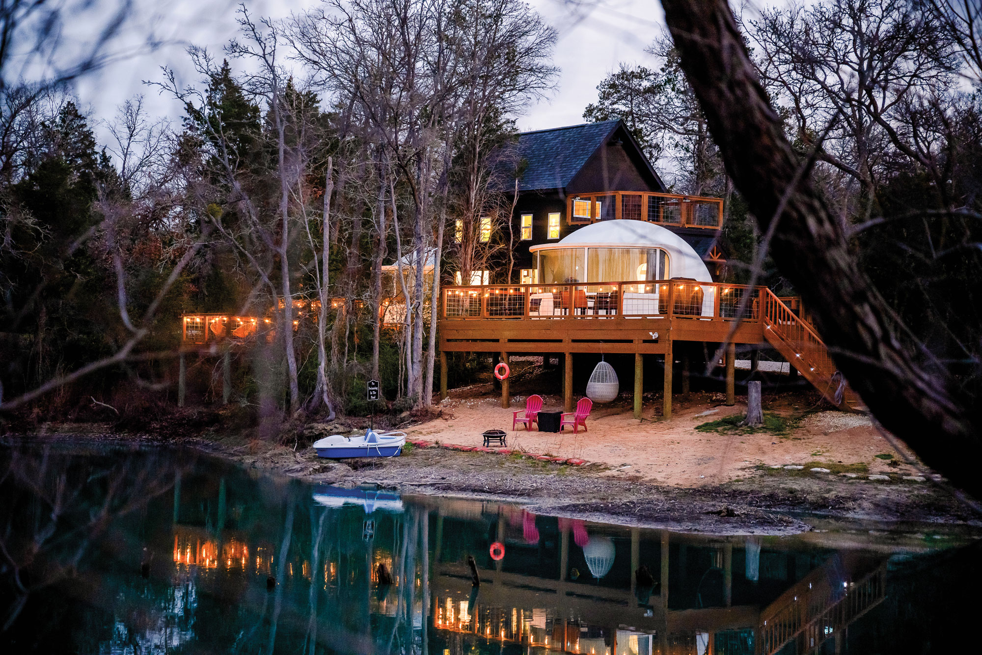 A house in front of a pond with a blue boat and pink deck chairs out front