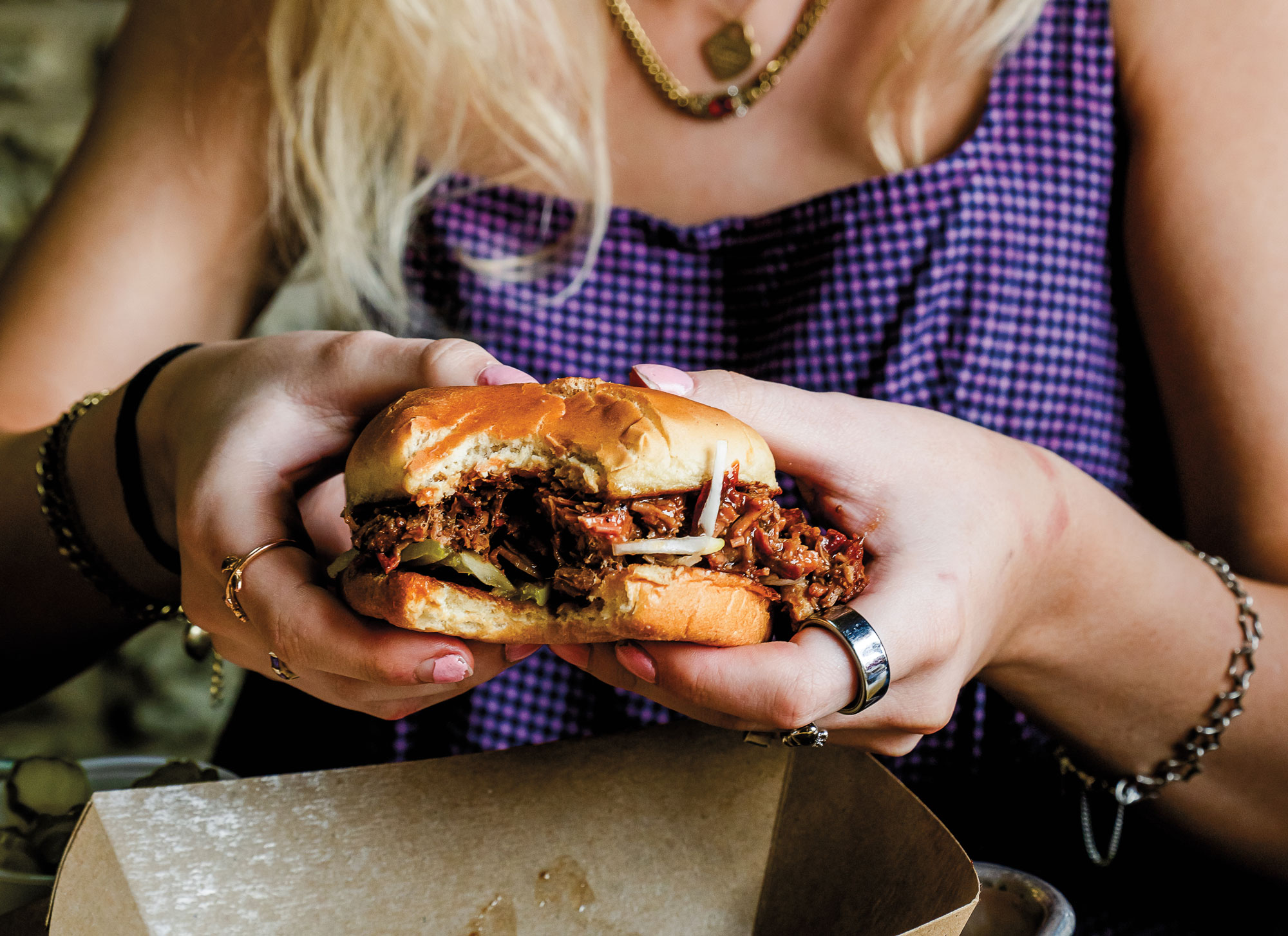 A blonde woman in a purple dress holding a chopped meat sandwich in a hamburger bun