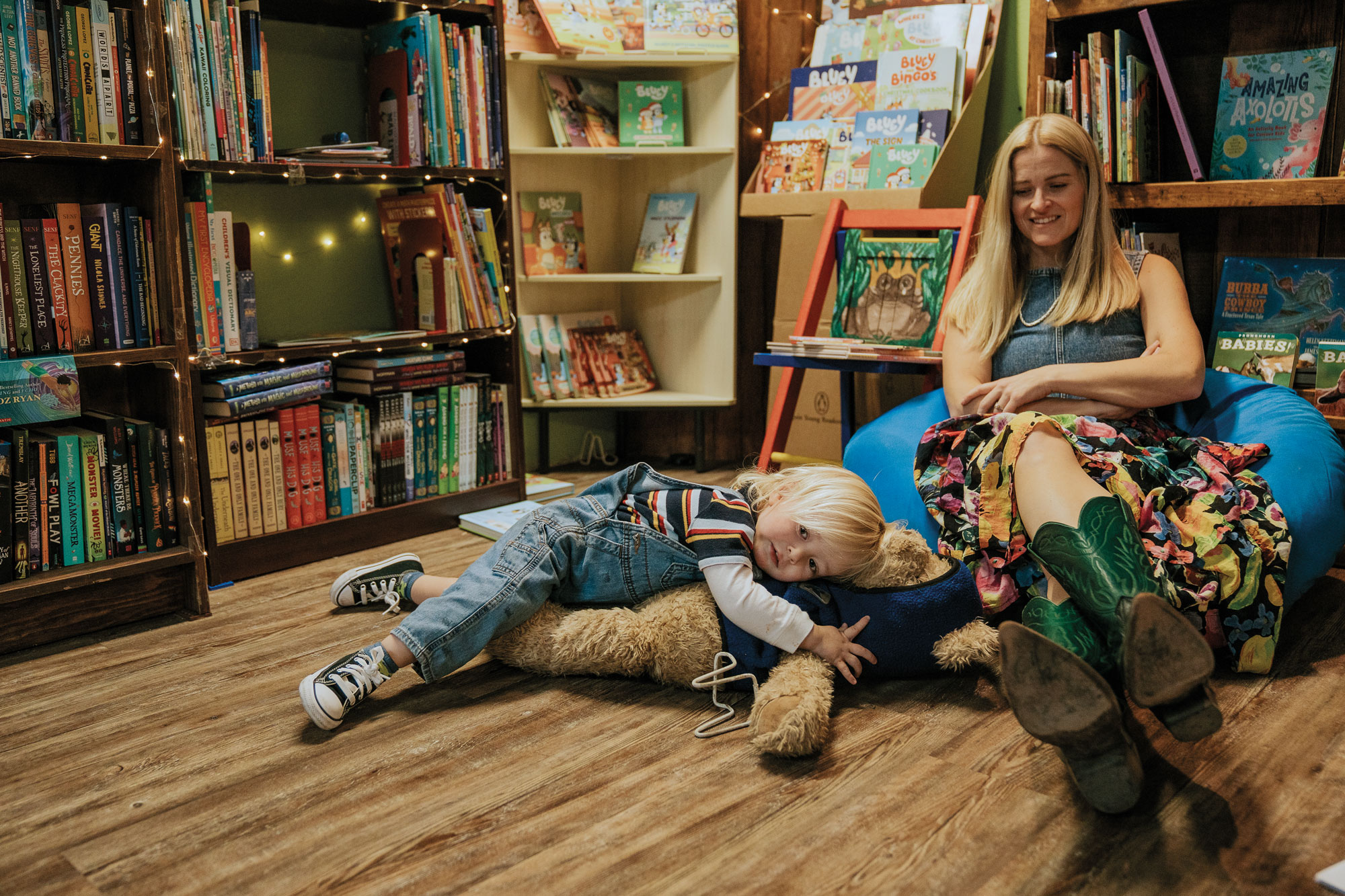 A blonde woman in her 30s sits on a blue beanbag chair while smiling at a small blonde boy thrown on the floor on top of a large teddy bear while in a bookstore.