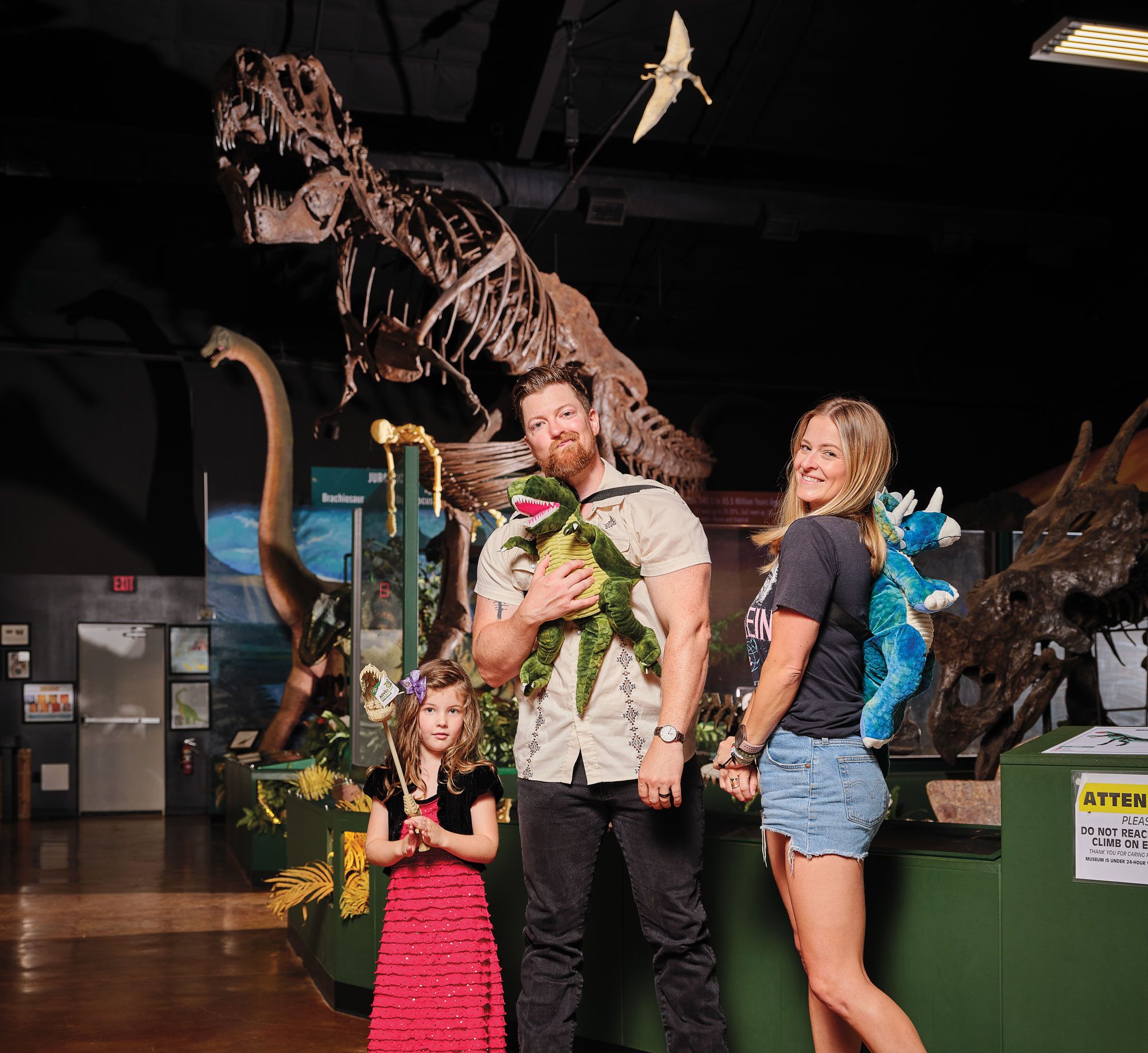 A man, woman, and their two young children—a daughter and a son—pose in a museum with a dinosaur skeleton behind them.