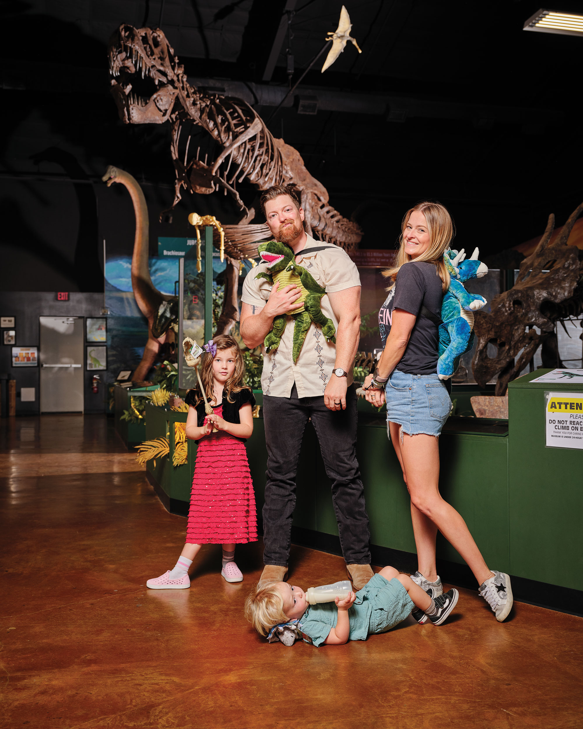 A man, woman, and their two young children—a daughter and a son—pose in a museum with a dinosaur skeleton behind them.