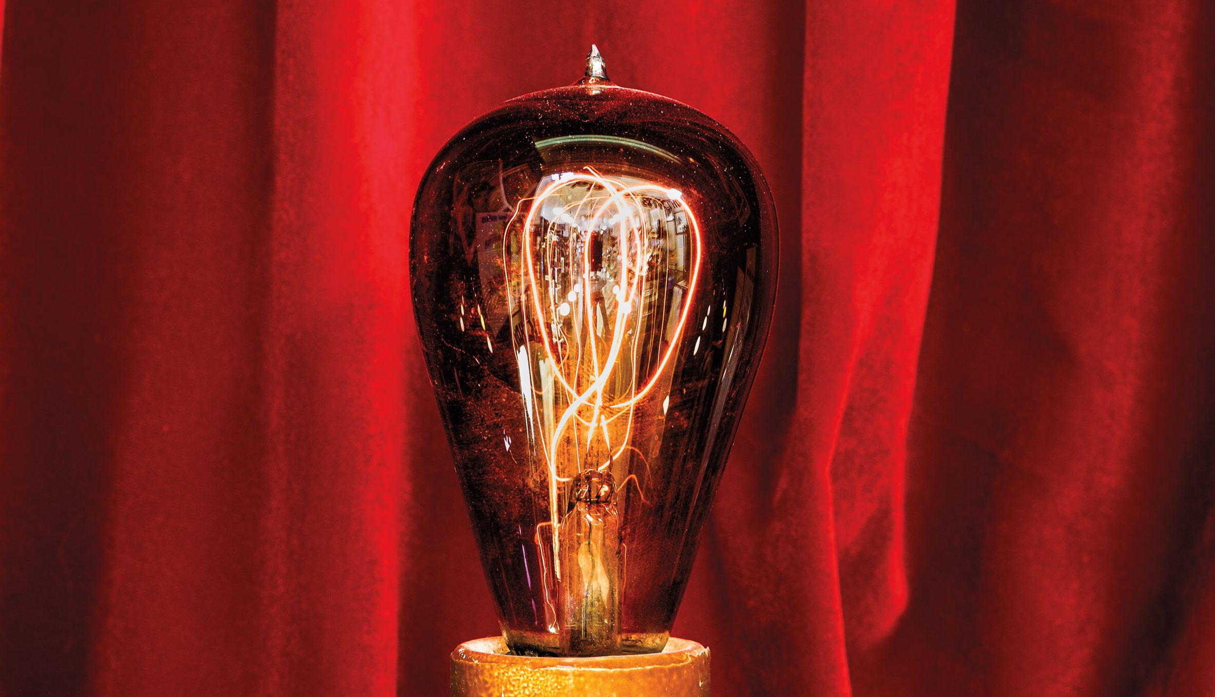 A close-up image of a lit lightbulb with orange wires inside in front of a red curtain