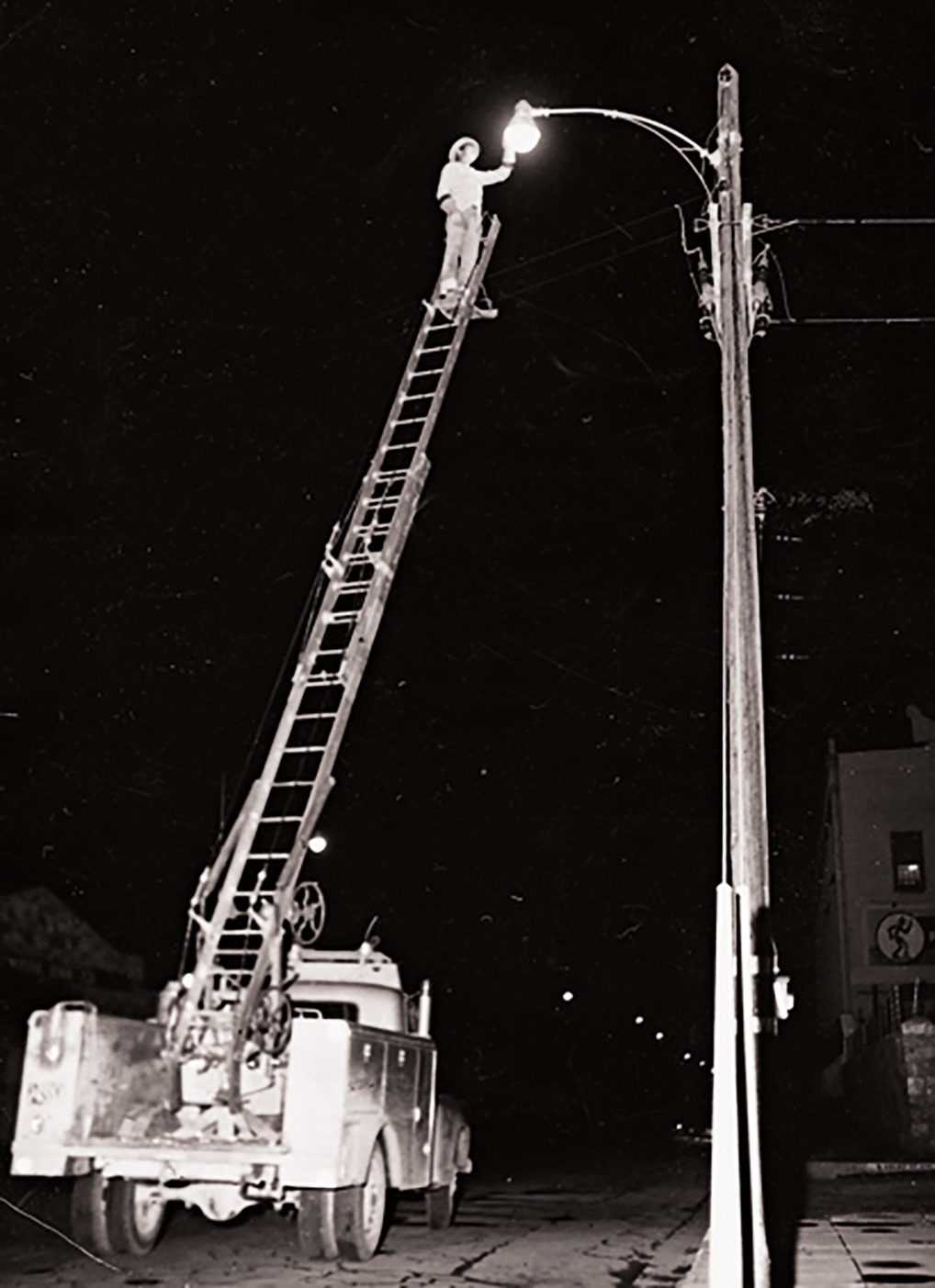 A man stands no a tall work ladder at night switching out a street light bulb