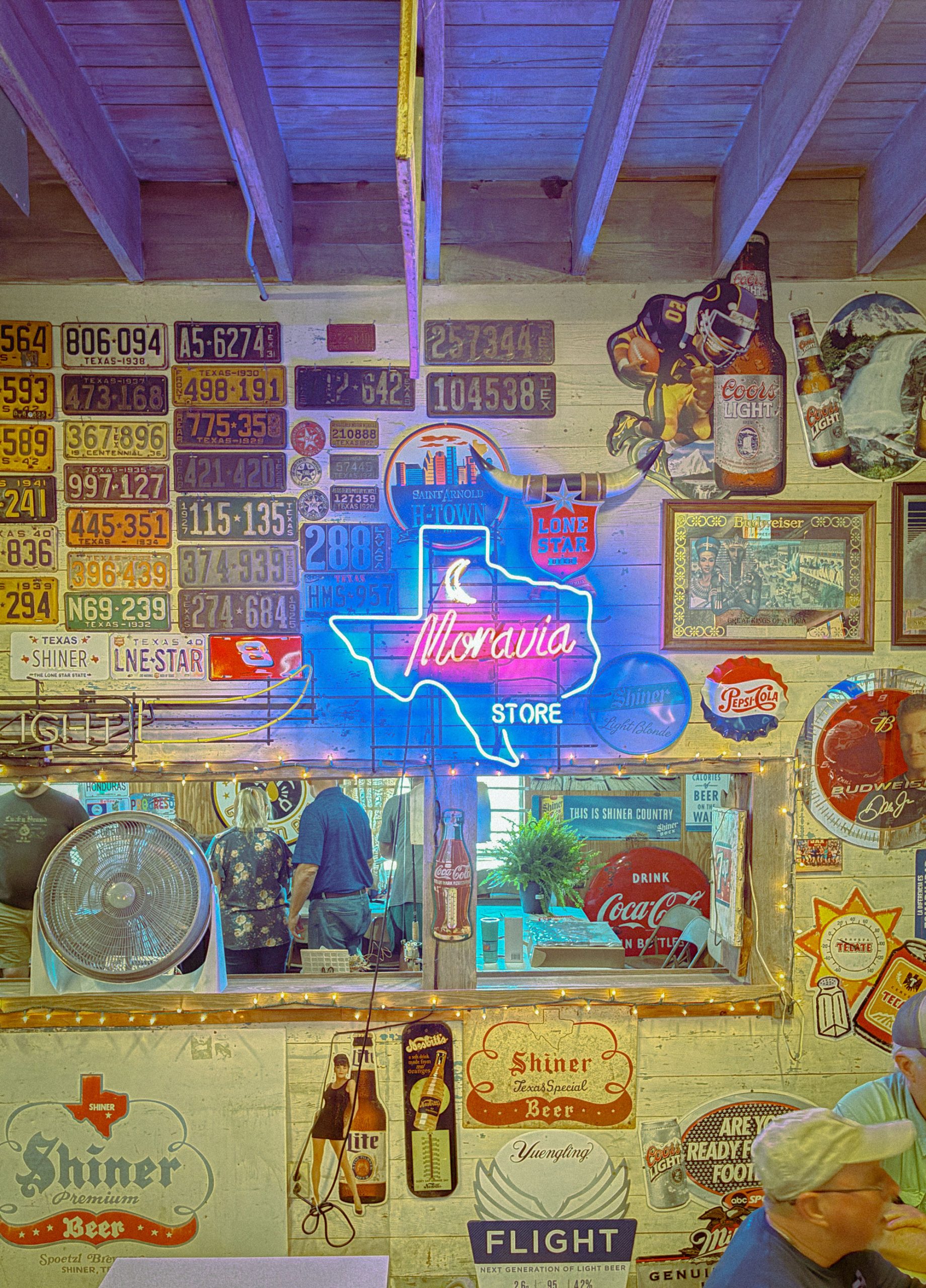 A neon sign in the shape of Texas reads "Moravia" and is hanging on a wall of a bar full of kitschy bar decor