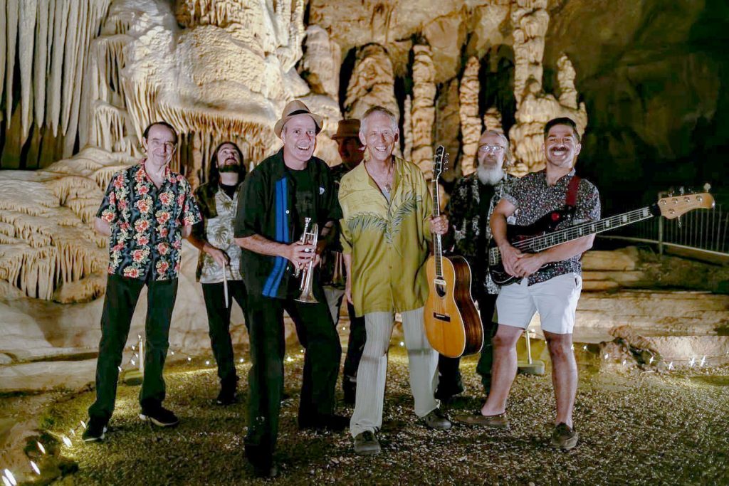 Six men, some holding a trumpet, guitar, and bass, pose inside a beige cavern