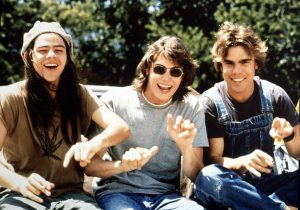 Three men dressed in 1970s attire sit and laugh in the sunlight
