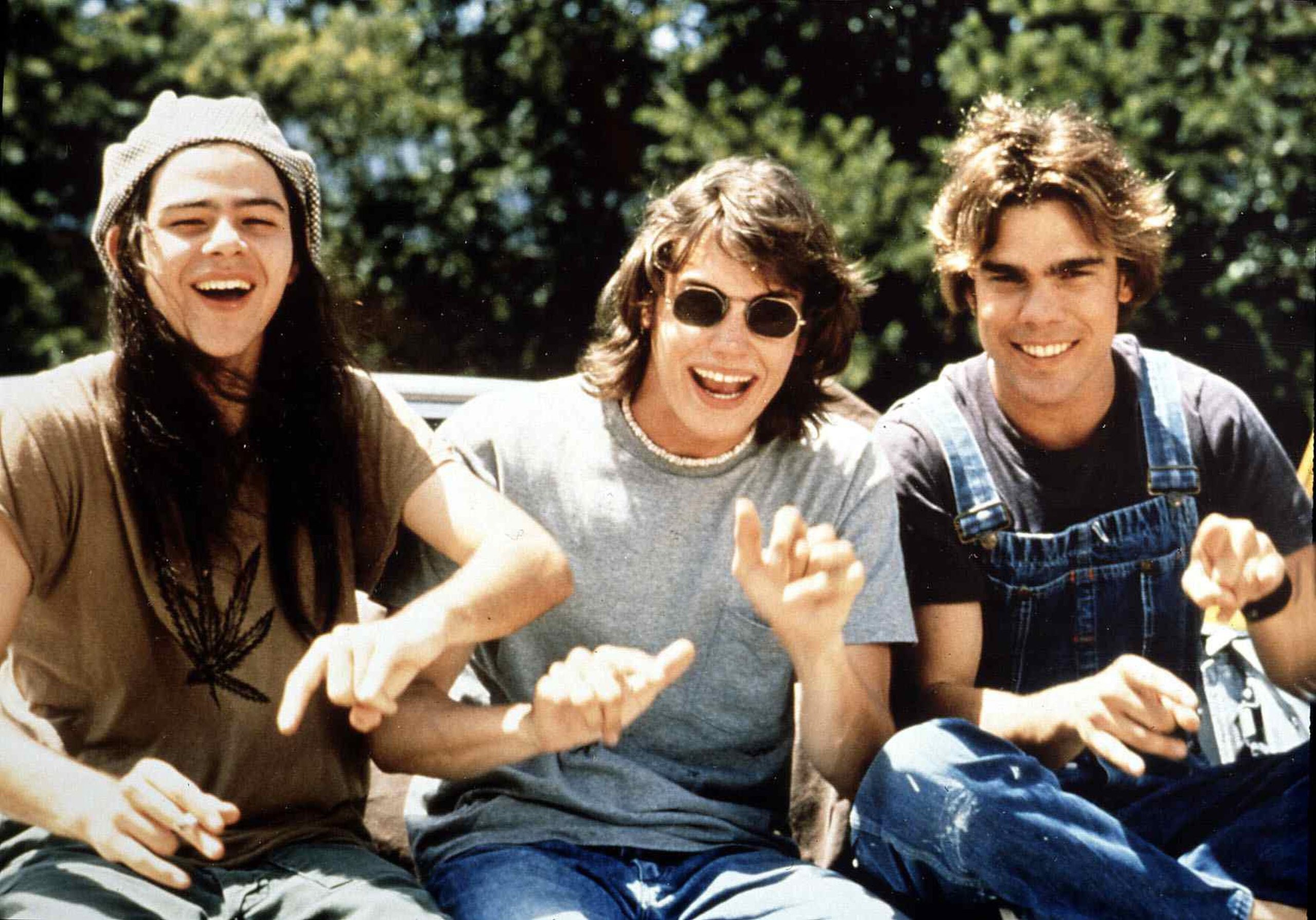 Three men dressed in 1970s attire sit and laugh in the sunlight