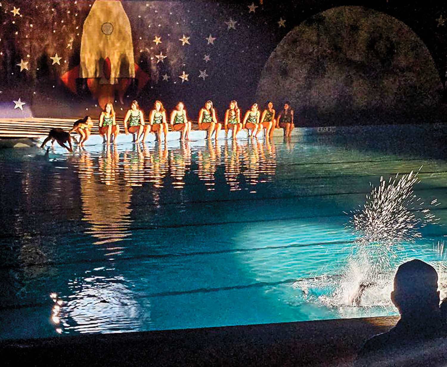 10 swimmers on the edge of a pool, one jumping in with a painting of a rocket behind them
