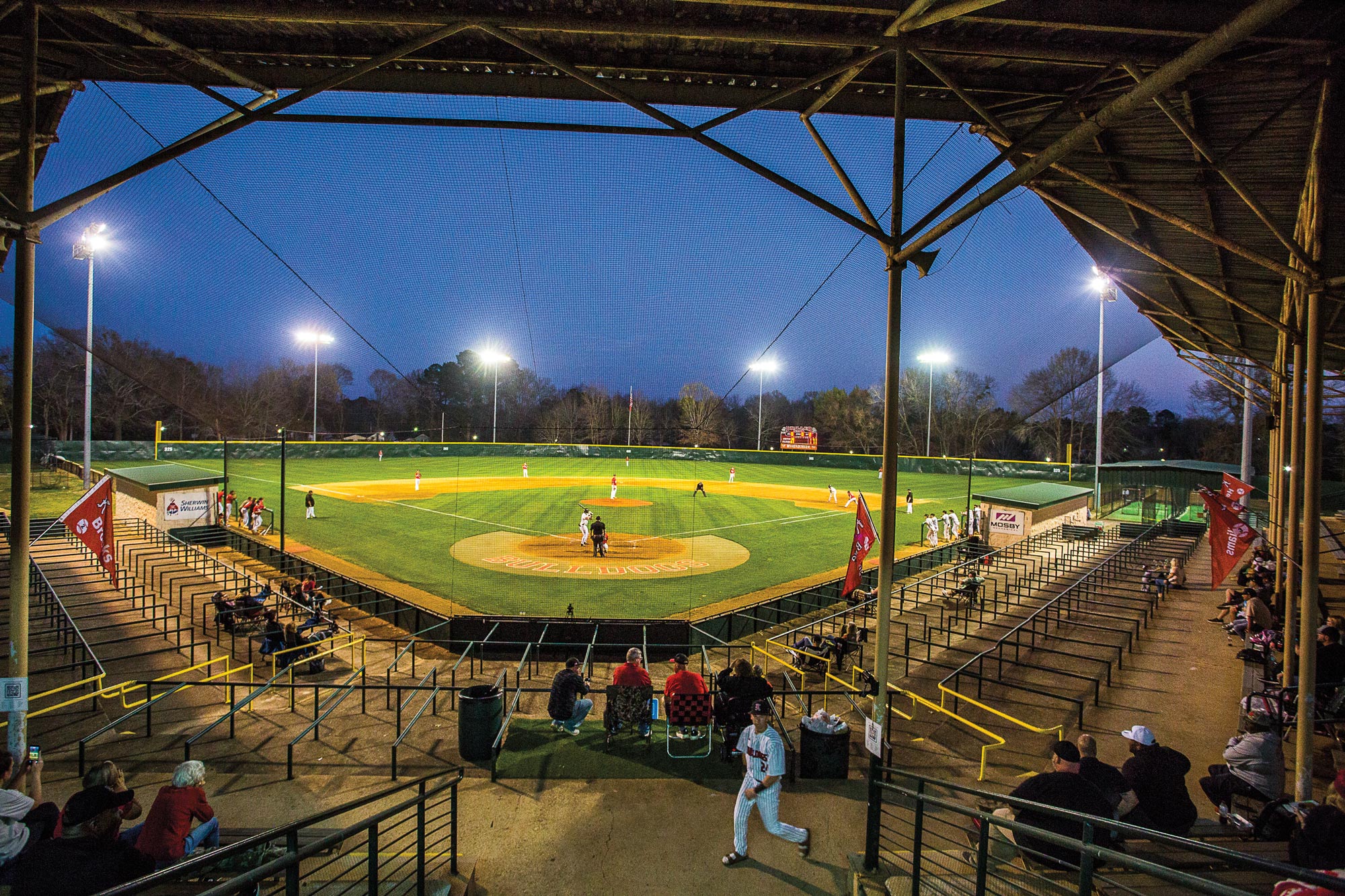 A baseball field at night during a game, with a person in full uniform walking in the foreground