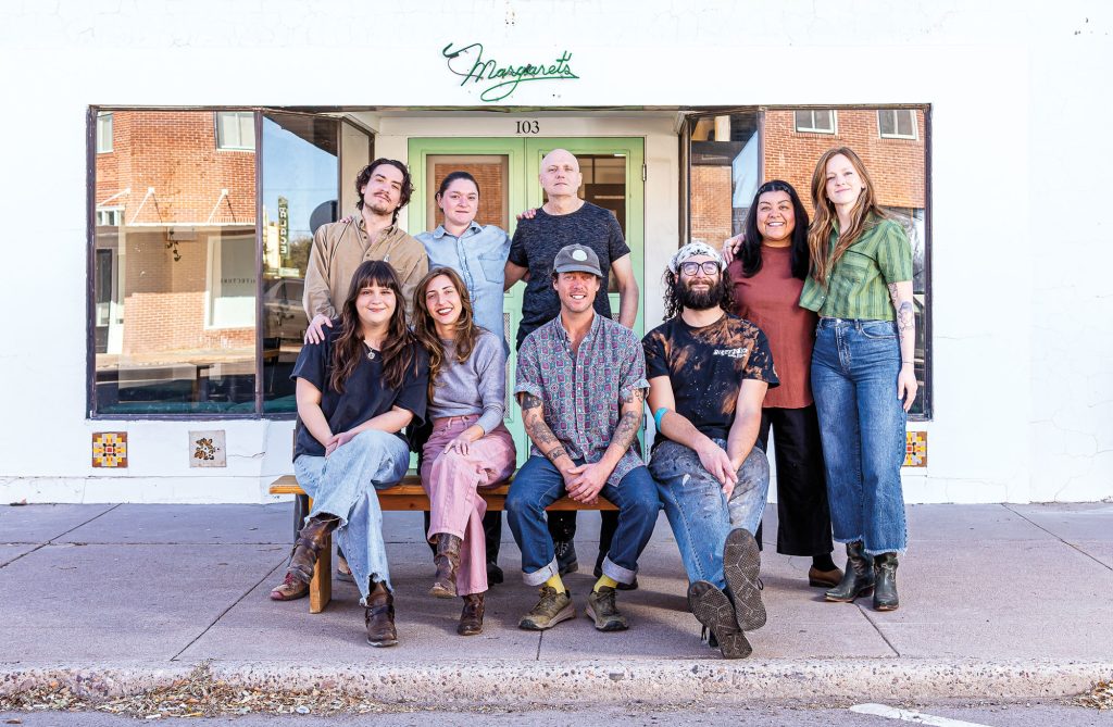 Nine people posed, four sitting, in front of a white building with a neon green sign that reads "Margaret's"