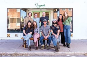 Nine people posed, four sitting, in front of a white building with a neon green sign that reads "Margaret's"