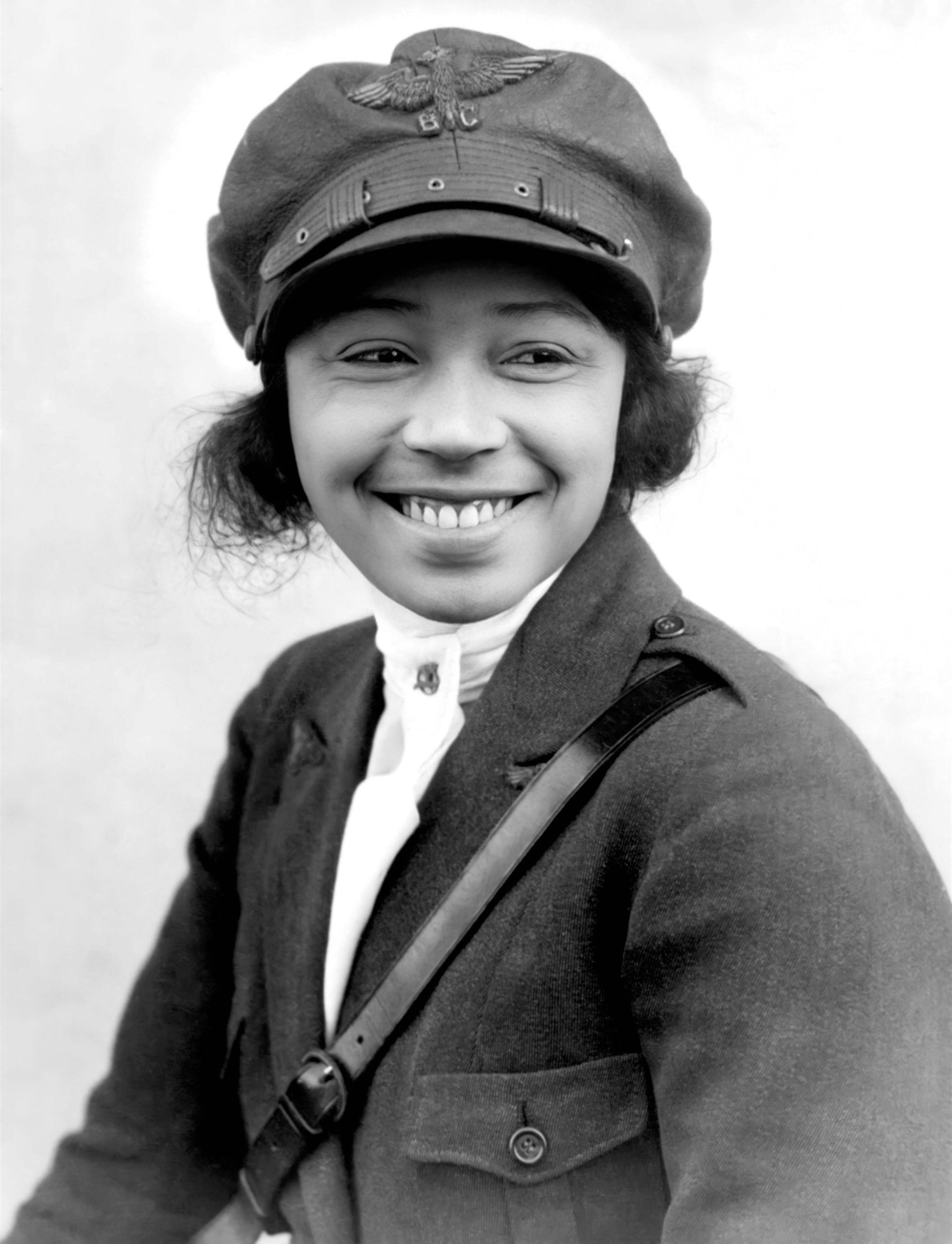 A smiling woman in a pilot's cap and jacket with a leather strap going down diagonally from her shoulder