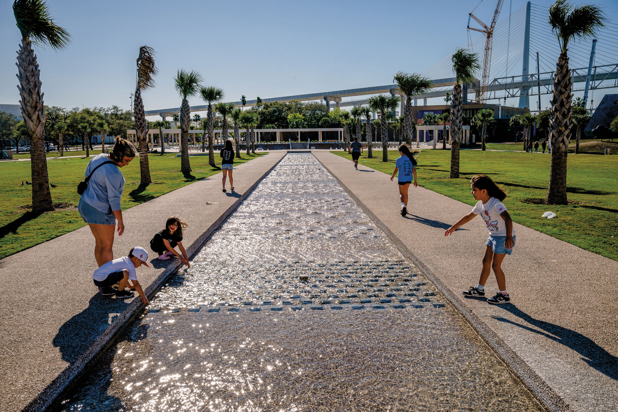 Children dipping their hands in a long, shallow pool of water in a walkway lined by palm trees