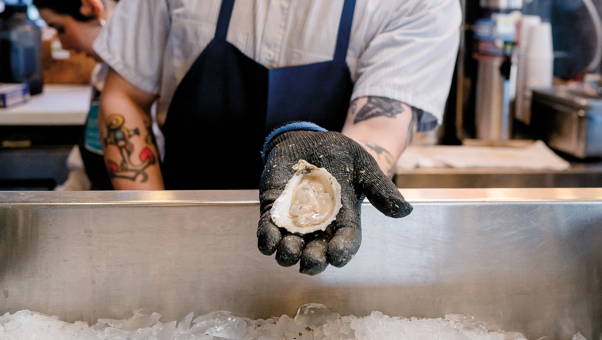 A gloved hand holding an oyster over a large tub of ice