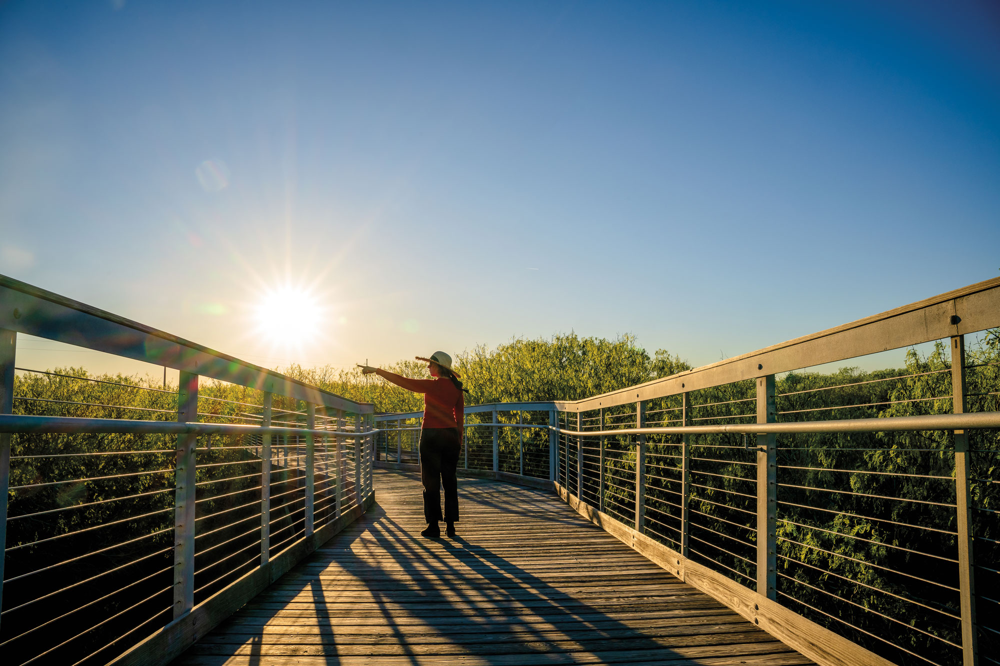 A person standing and pointing on a wooden bridge with the sun low on the horizon