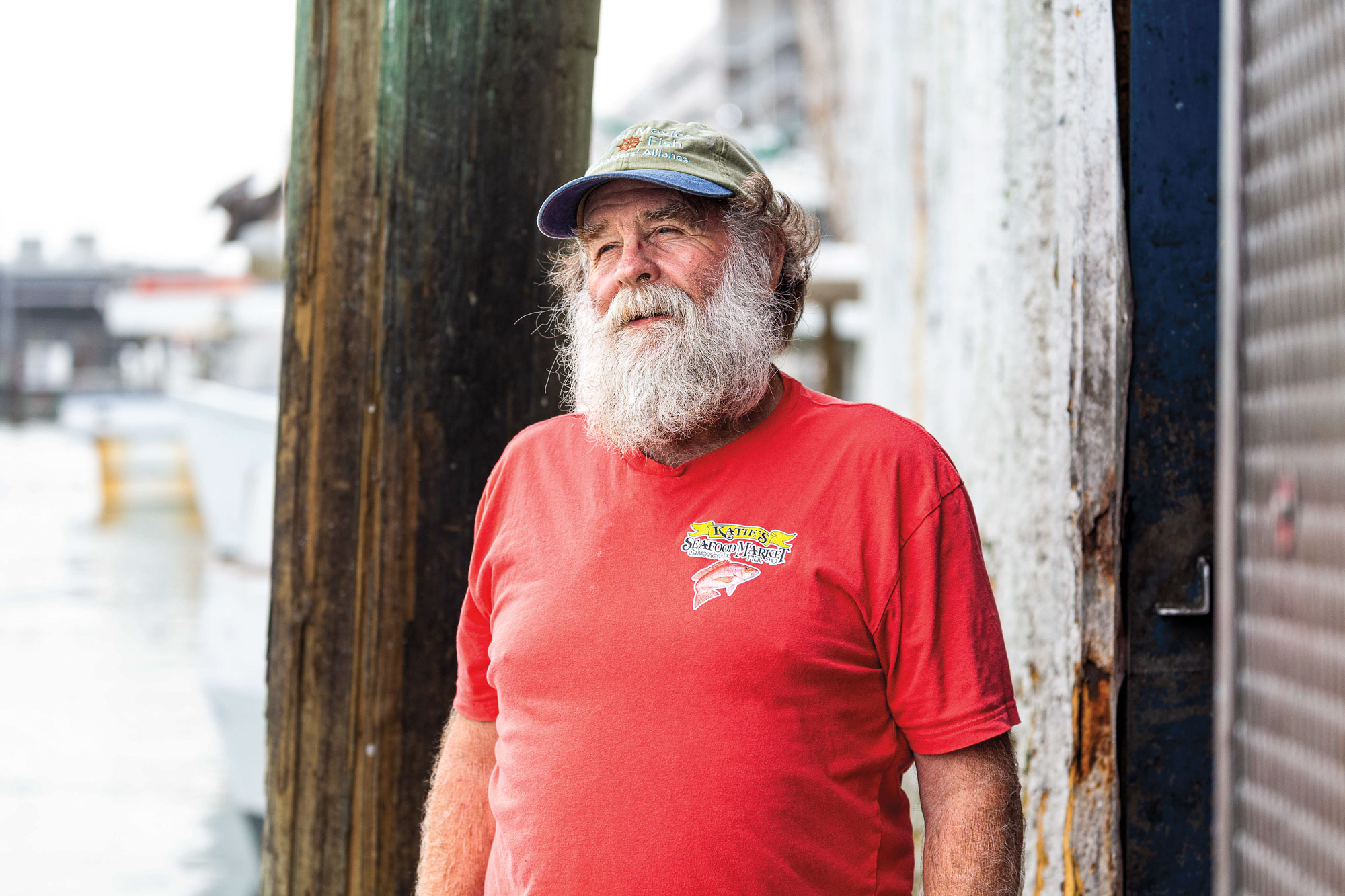 A man with a long, white beard wearing a baseball cap and a red T-shirt that reads Katie's Seafood Market