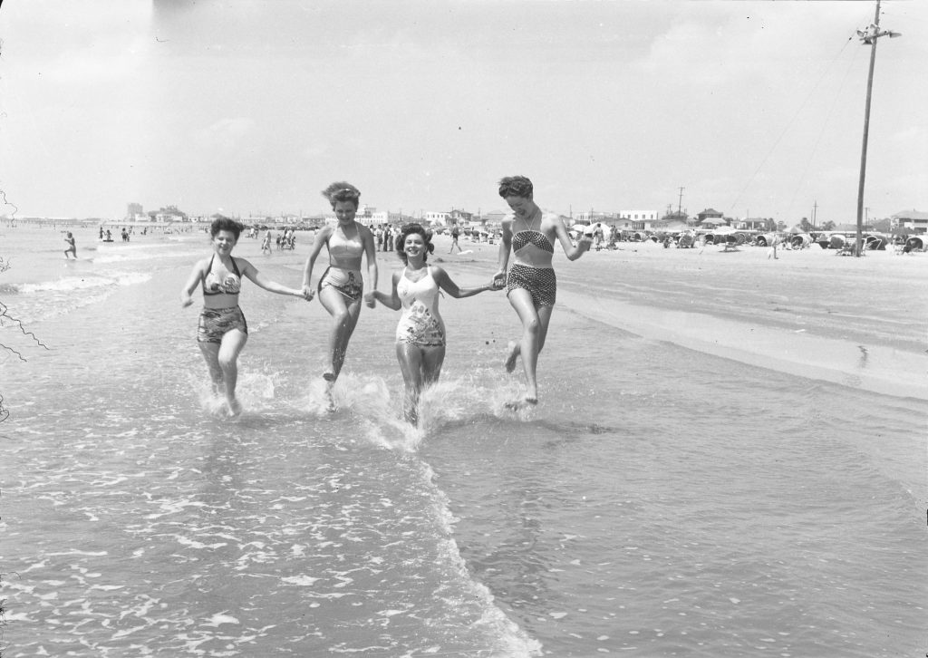 Four women, hand-in-hand running through shallow water on a beach with many people behind them