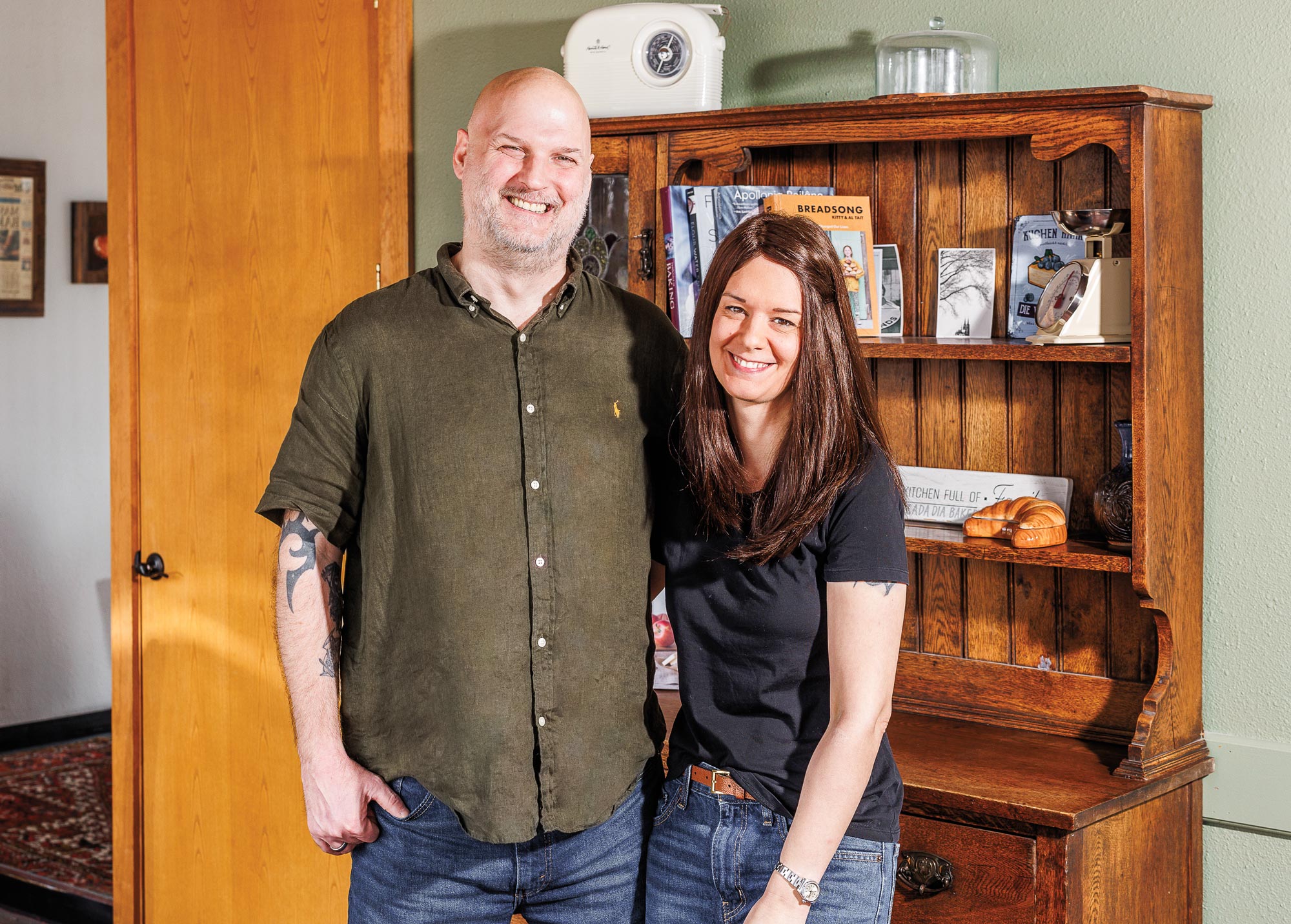A man in a green button-down shirt and jeans next to a woman in a black T-shirt and jeans in front of a wooden hutch with books and other items on it