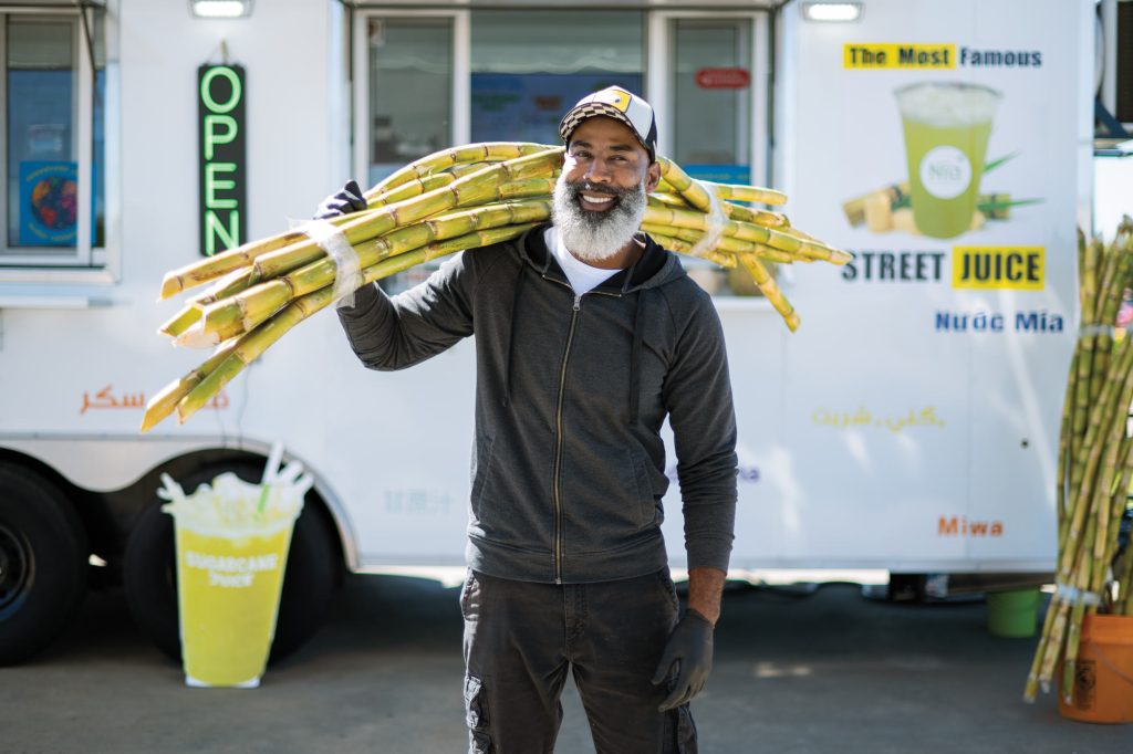 A man with a white beard and a baseball cap on holds a big bundle of green sugarcane on his shoulder