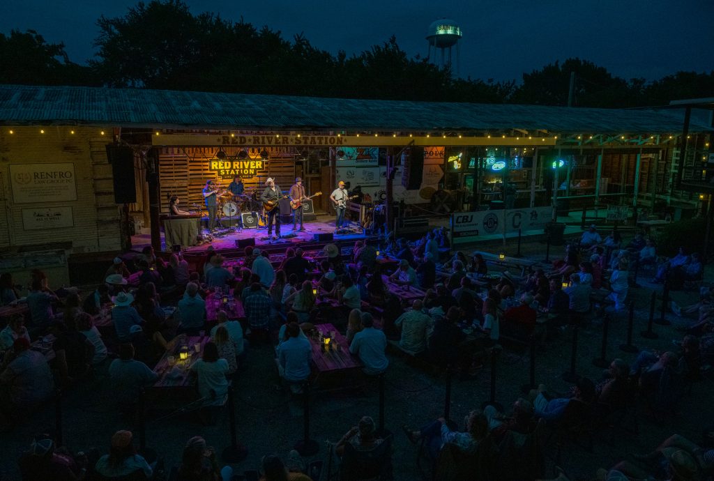 People sitting in chairs in front of a large stage with six musicians playing under and in front of multiple signs that say Red River Station at night