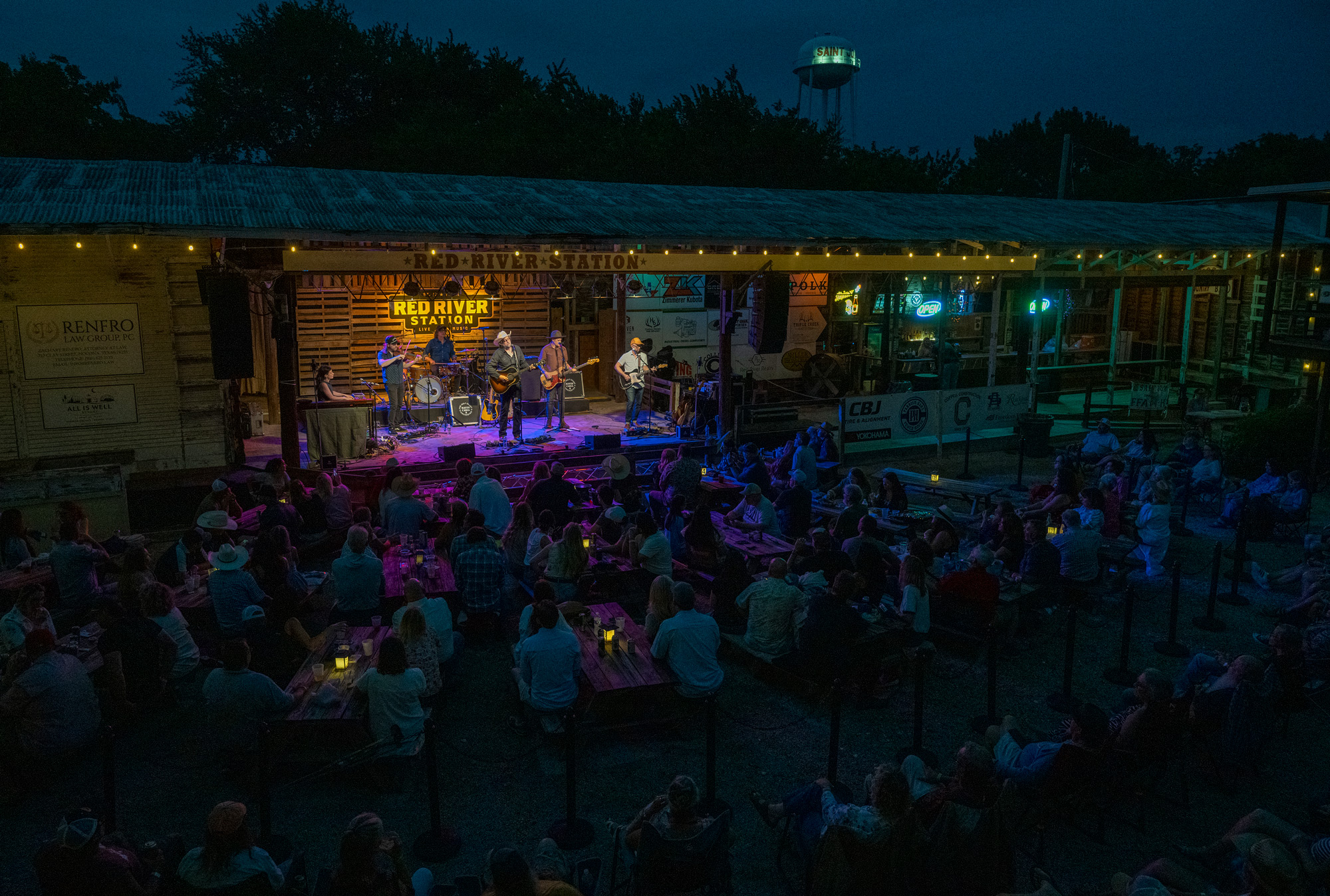 People sitting in chairs in front of a large stage with six musicians playing under and in front of multiple signs that say Red River Station at night