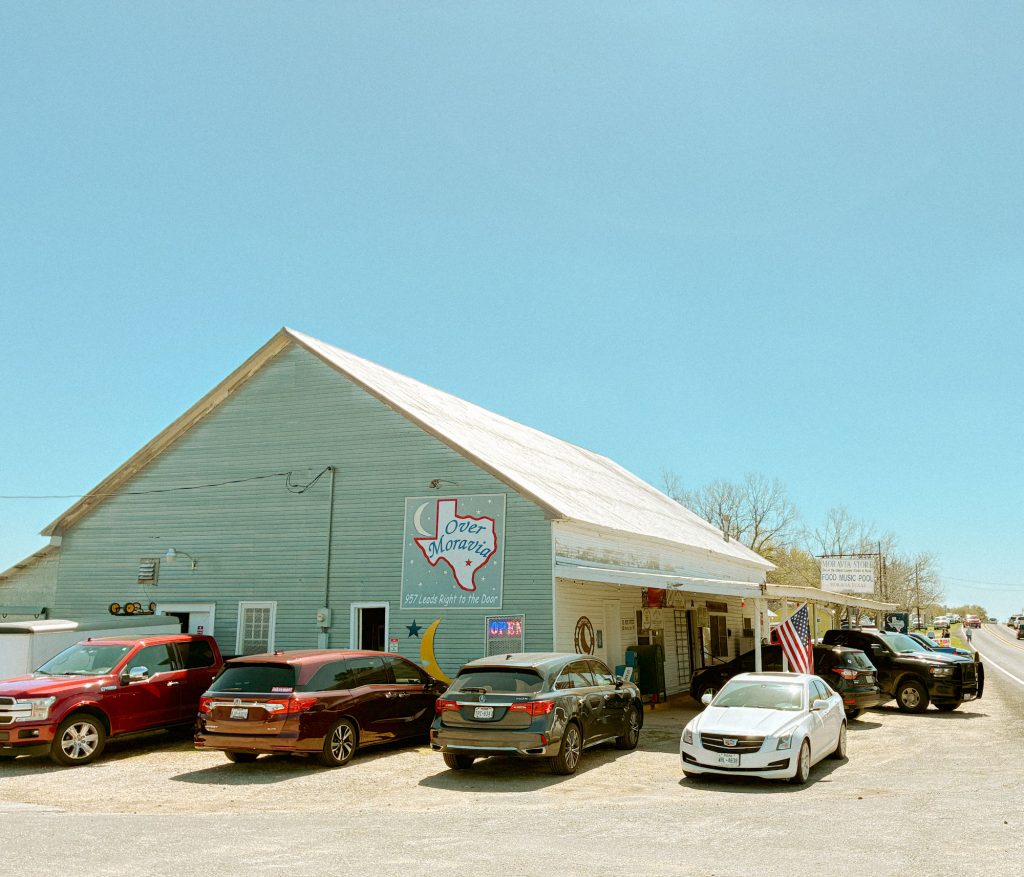 A building made of wooden slats, cars parked around it. A sign in the shape of Texas hangs on the side and reads "Moravia"