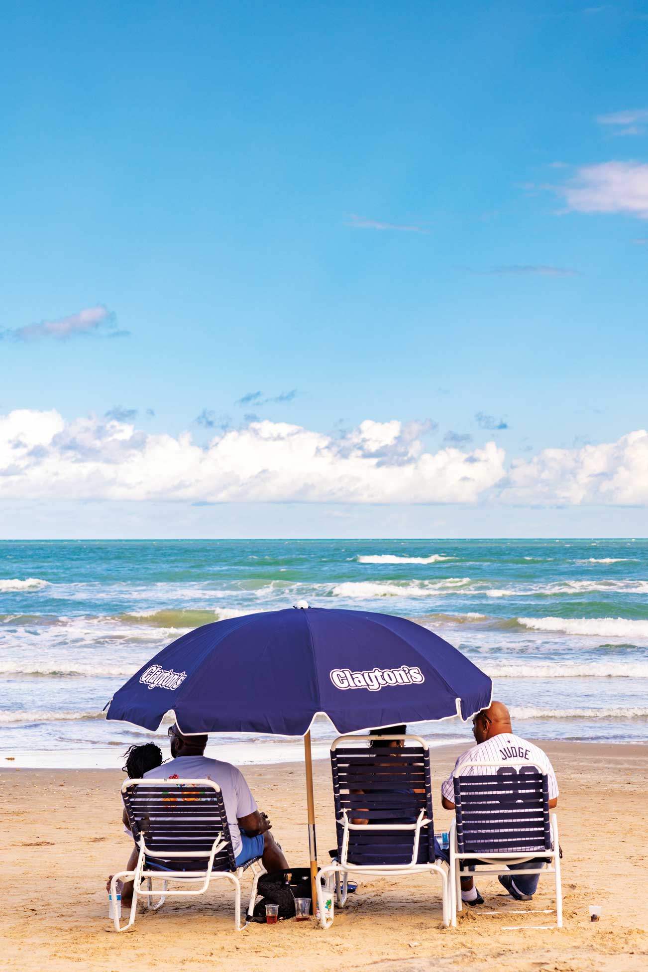 People sit in beach chairs facing the ocean under an umbrella that reads Clatyon's