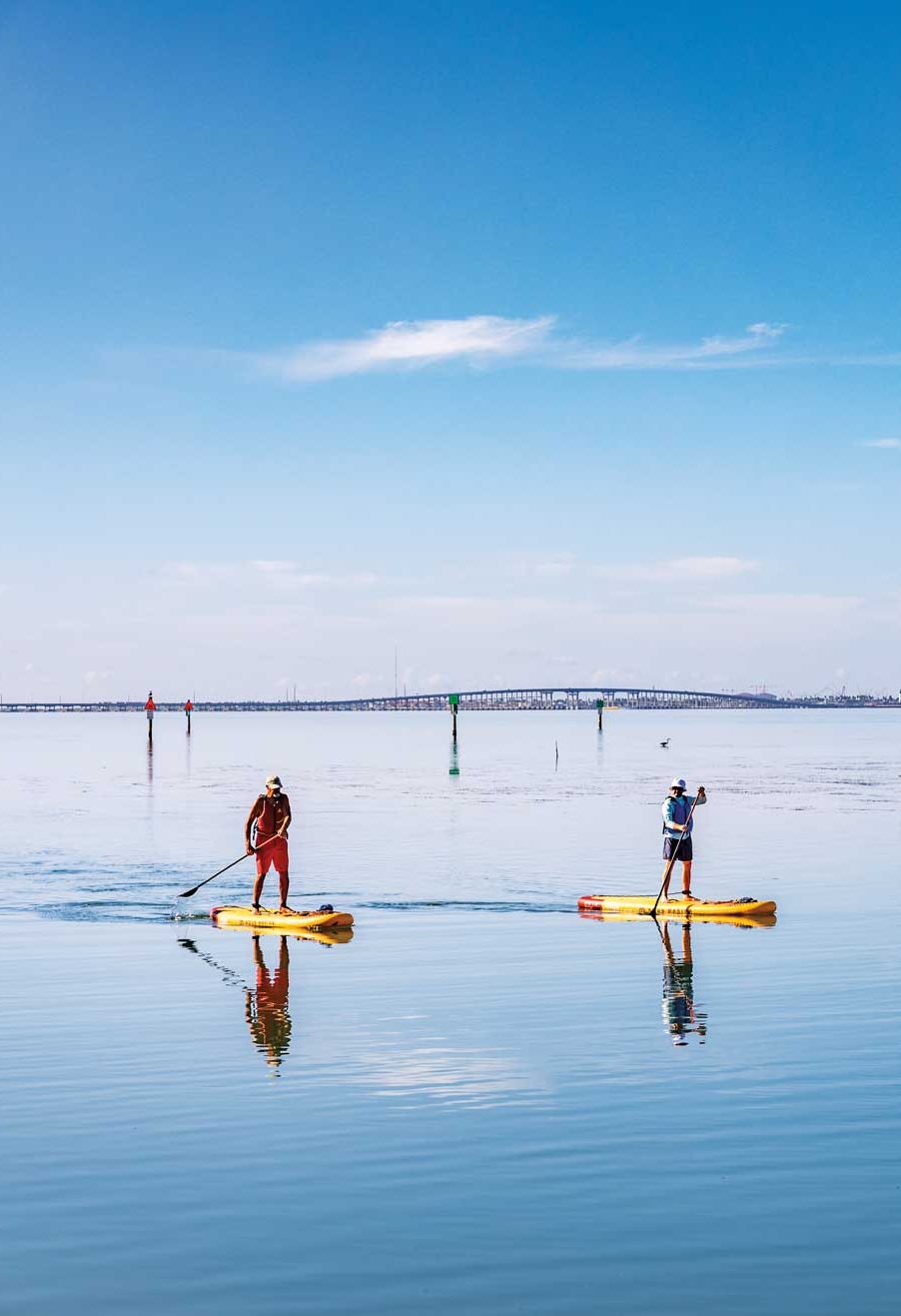 Two people on paddleboards on a big blue body of water with a bridge off in the far distance